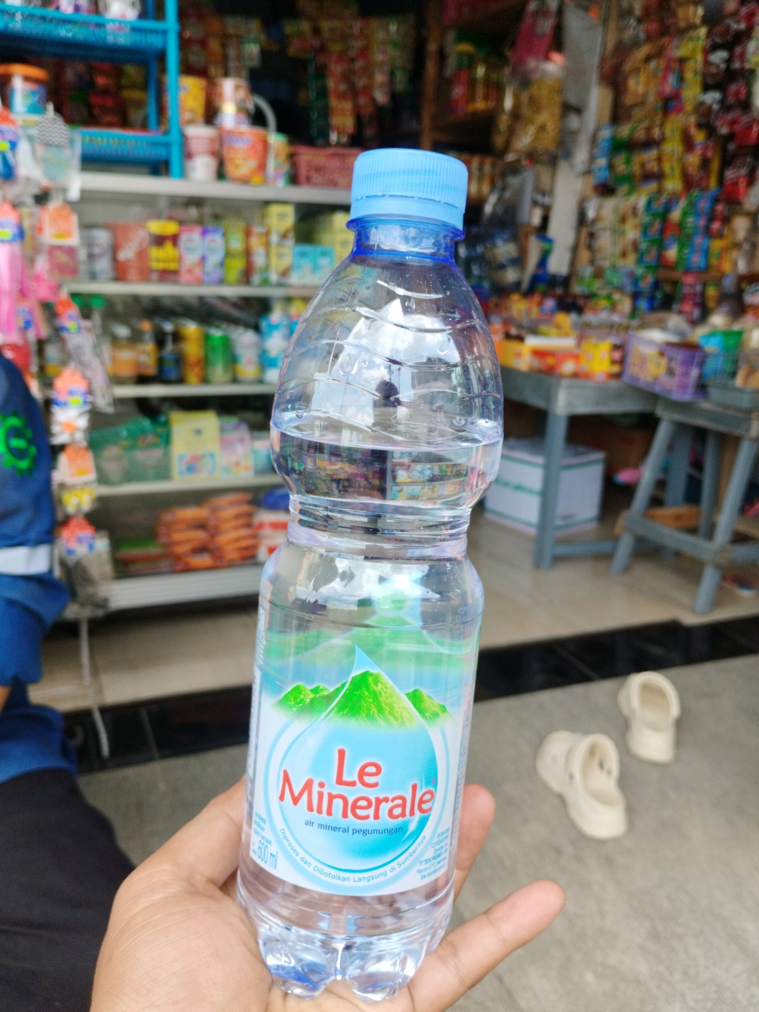 A person's hand is holding a clear plastic bottle of "Le Minerale" bottled water. The bottle has a blue cap and features a label with green mountains and the brand name. The background suggests a retail environment, possibly a small shop or convenience store in Pakuncen, Indonesia. Shelves filled with various packaged goods, including snacks and drinks, are visible. In the lower right corner of the image, two light-colored clogs are placed on the ground. The lighting is natural, and the focus is on the water bottle.