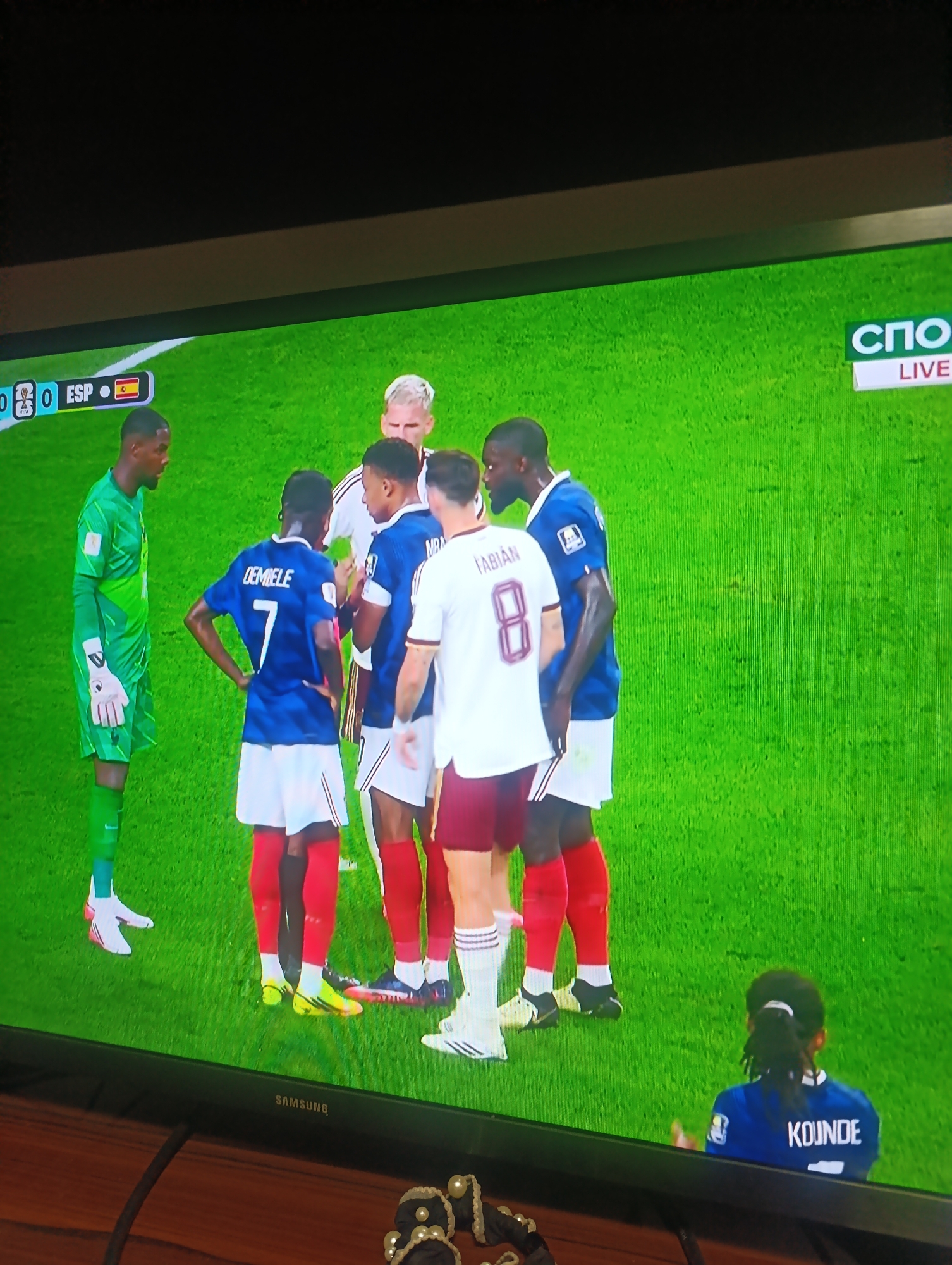A television screen displays a soccer match. On the left, a soccer player in a green uniform with white gloves stands facing right. In the center, four soccer players in blue uniforms with white shorts and red socks huddle together. One player in the foreground wears a blue jersey with "DEMBELE 7" on the back. Another player in the center wears a white jersey with "FABIAN 8" on the back and red shorts. To the right, a player in a blue jersey with white shorts and red socks stands facing left, looking at the players in the center. In the lower right corner, a person with dark hair in a blue uniform faces away from the camera, looking towards the match. The background is a green soccer field. A score graphic in the upper left corner shows "0 - 0 ESP" with a Spanish flag. A broadcast logo in the upper right corner shows "СПО LIVE". The television is placed on a wooden surface.