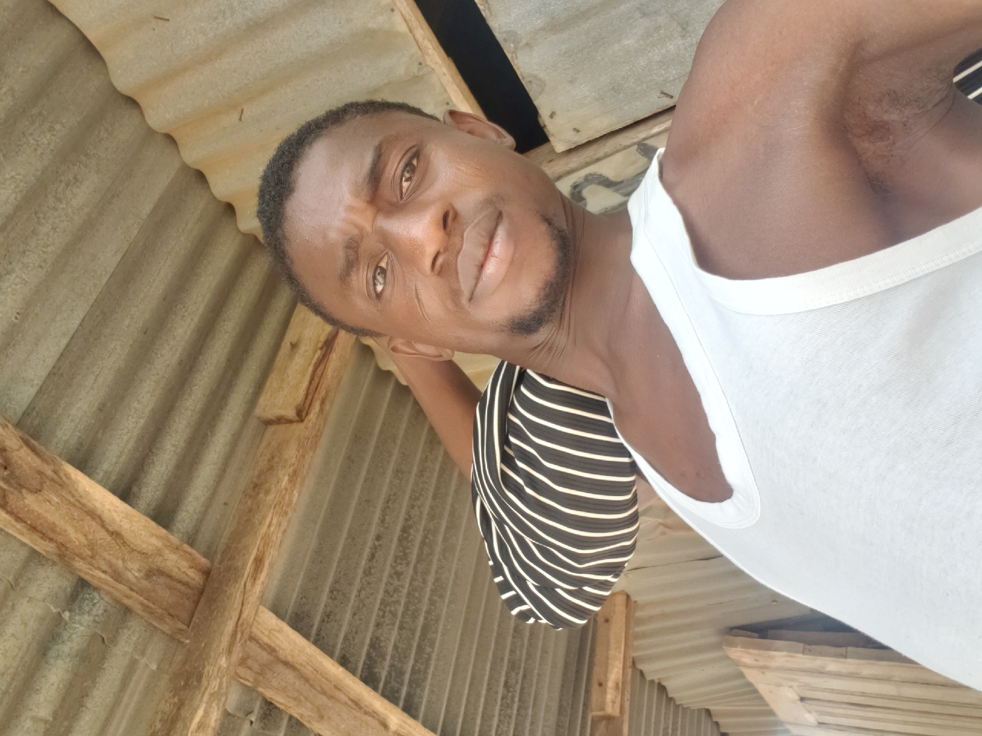 A close-up, diagonally oriented image captures a man looking directly at the camera. He has dark skin, short dark hair, and a short beard or stubble. His expression is serious or neutral. He is wearing a white sleeveless undershirt. A black and white striped garment is visible near his left arm, possibly held in his hand.
The background shows an overhead structure, likely a ceiling or roof. It consists of corrugated metal sheets, which appear weathered and light-colored, possibly grey or tan. These sheets are intersected by rough-hewn wooden beams or supports, forming a crisscross pattern. The lighting suggests it is daytime, with bright, even illumination. The structure appears rustic and functional. No visible text is present in the image. The setting, with its corrugated roof and wooden framework, aligns with common building constructions found in various regions, including Adankolo, Nigeria.
