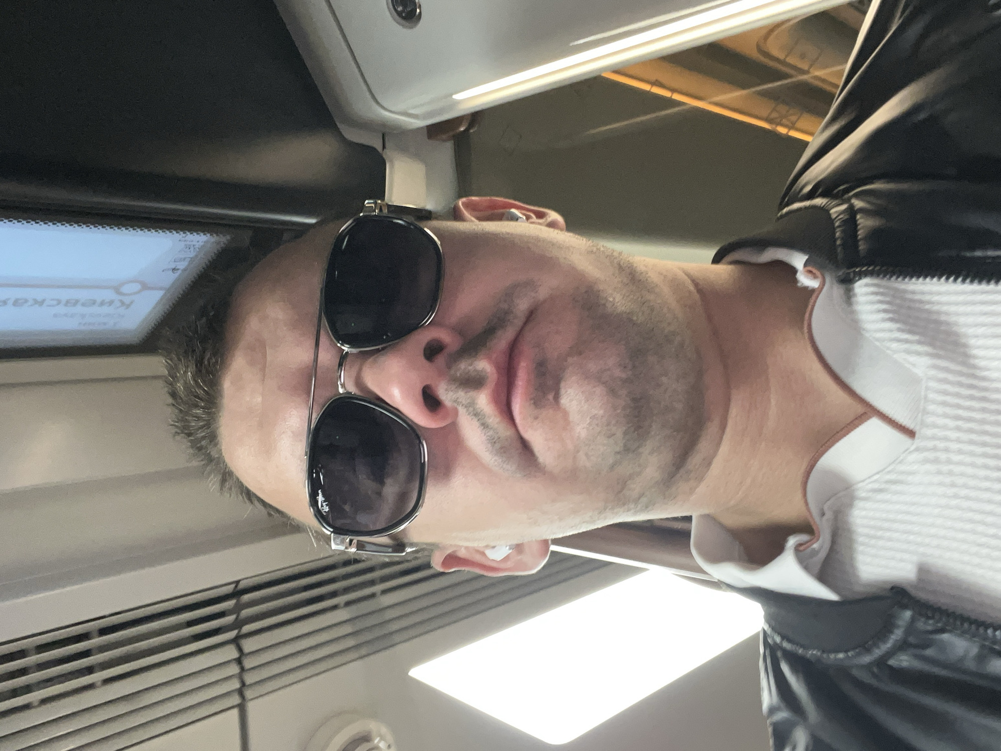 A low-angle, medium shot captures a man wearing sunglasses, a white collared shirt, and a black jacket. He is likely in a subway car in Moscow, Russia, given the partial view of Cyrillic text on a digital display in the background that reads "Киевская," referring to the Kievskaya metro station. The man appears to be looking directly at the camera. He has earbuds in both ears. The lighting in the scene is somewhat dim, with a bright light source from the left.