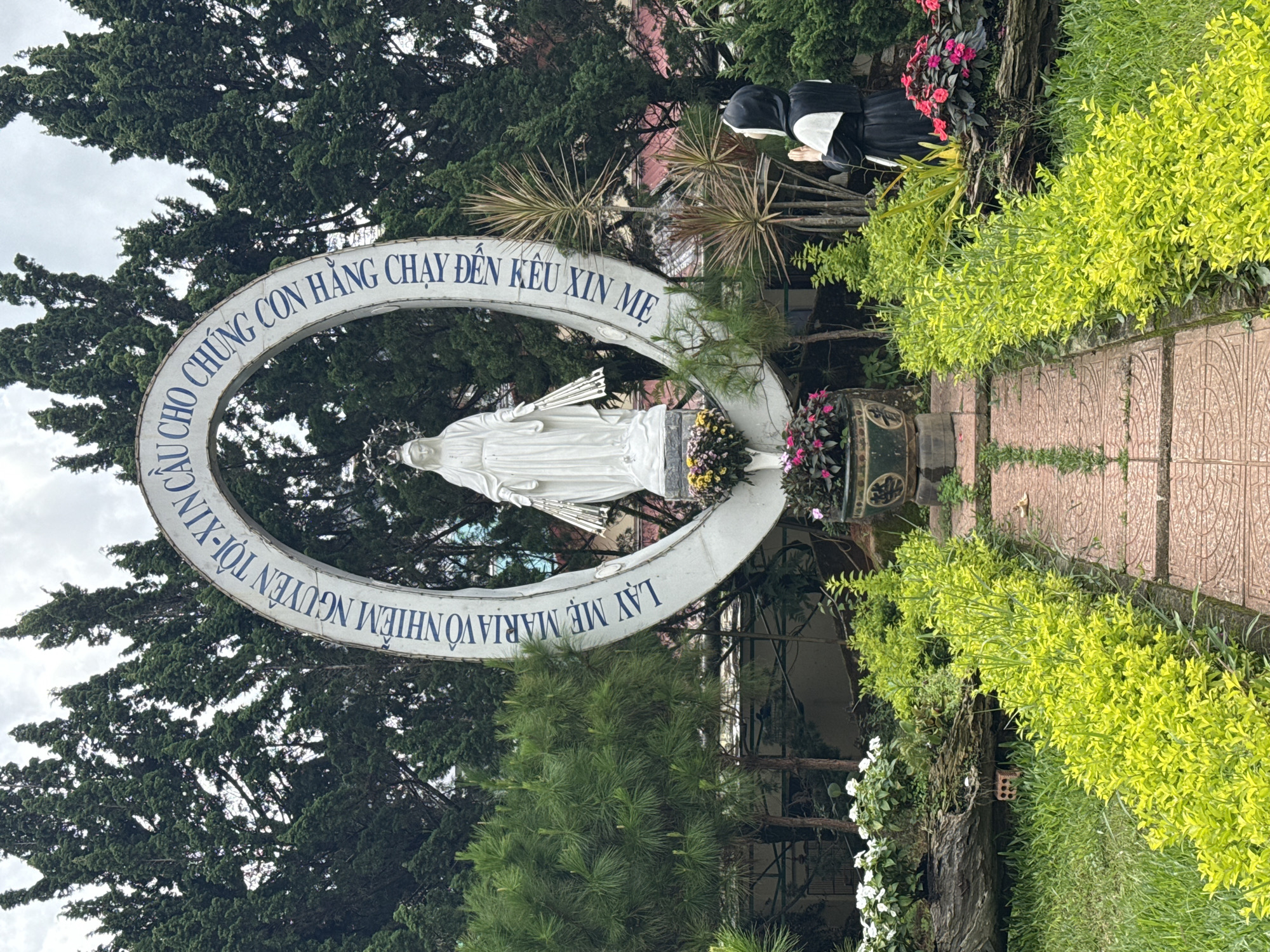 This image depicts an outdoor Marian shrine, likely located in Da Lat, Vietnam, given the cultural context and location hint. At the heart of the scene is a white statue of the Virgin Mary, standing within a large, circular or arched white structure. The arch is adorned with blue Vietnamese text, which reads: "LẠY MẸ MARIA VÔ NHIỄM NGUYÊN TỘI-XIN CHO CHÚNG CON HẲNG CHẠY ĐẾN KÊU XIN MẸ". This translates to "O Mary conceived without sin, pray for us who have recourse to thee."
To the right of the Virgin Mary statue, a second statue portrays a kneeling figure, possibly a nun or a devotee, dressed in a black habit and white coif, positioned in an act of prayer or reverence. The shrine is surrounded by lush and varied foliage, including tall, dark green coniferous trees in the background, lighter green leafy bushes bordering the scene, and some palm-like plants. Potted flowers, displaying pink and yellow hues, are placed at the base of the statues, adding splashes of color. A paved pathway with patterned tiles is visible in the foreground, leading towards the shrine. The sky above appears overcast, indicating a cloudy daytime setting. The overall scene conveys a serene and devotional ambiance.