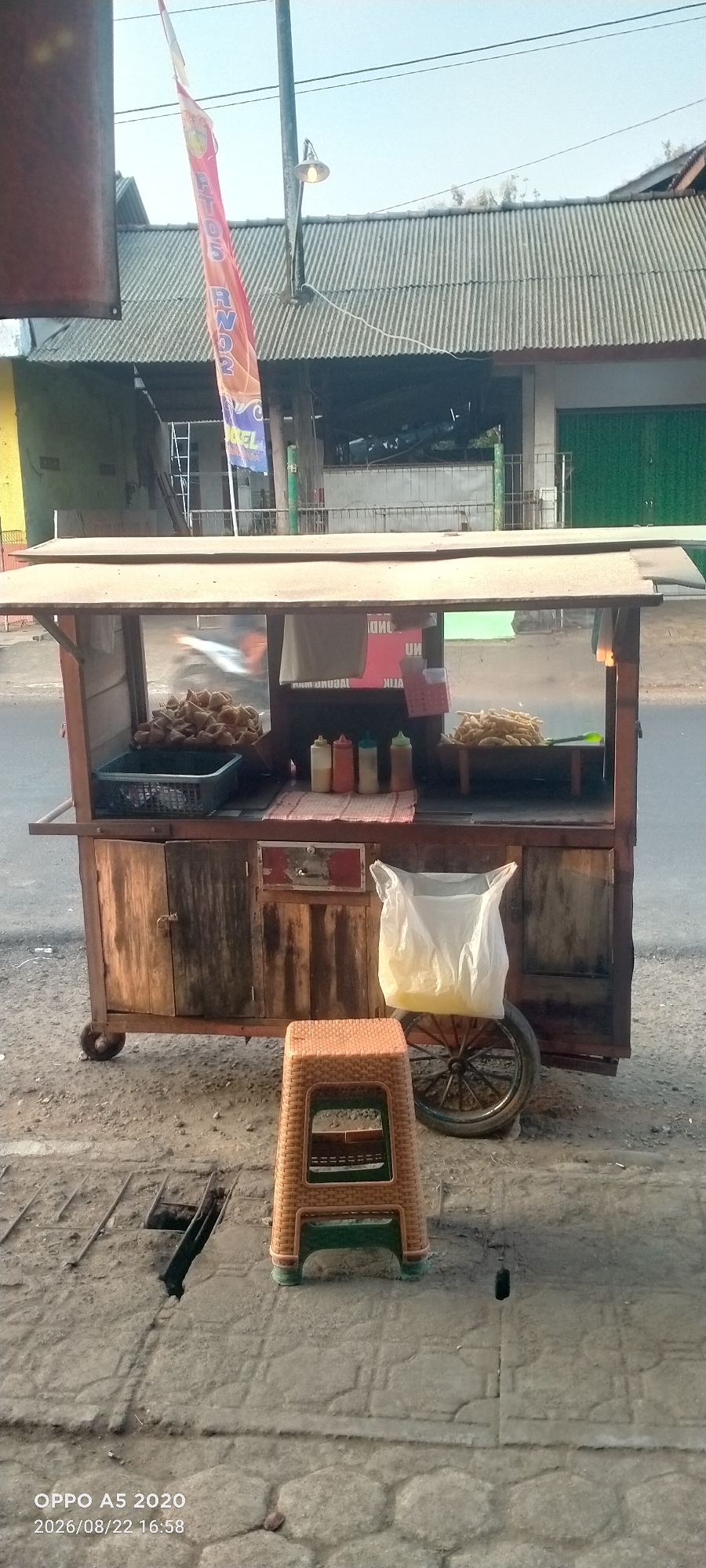 The image shows a food cart in Kedung, Indonesia. The cart is wooden with a metal frame and wheels. It is stocked with various food items, including what appear to be fried snacks and bottles of condiments. A plastic stool is placed in front of the cart. In the background, there is a building with a corrugated metal roof and a street with a few vehicles visible. A flag is also hanging from a pole. The image appears to have been taken during the daytime, with natural light illuminating the scene. The ground in front of the cart is paved with concrete. The overall impression is of a typical street food vendor setup in an urban or semi-urban environment.