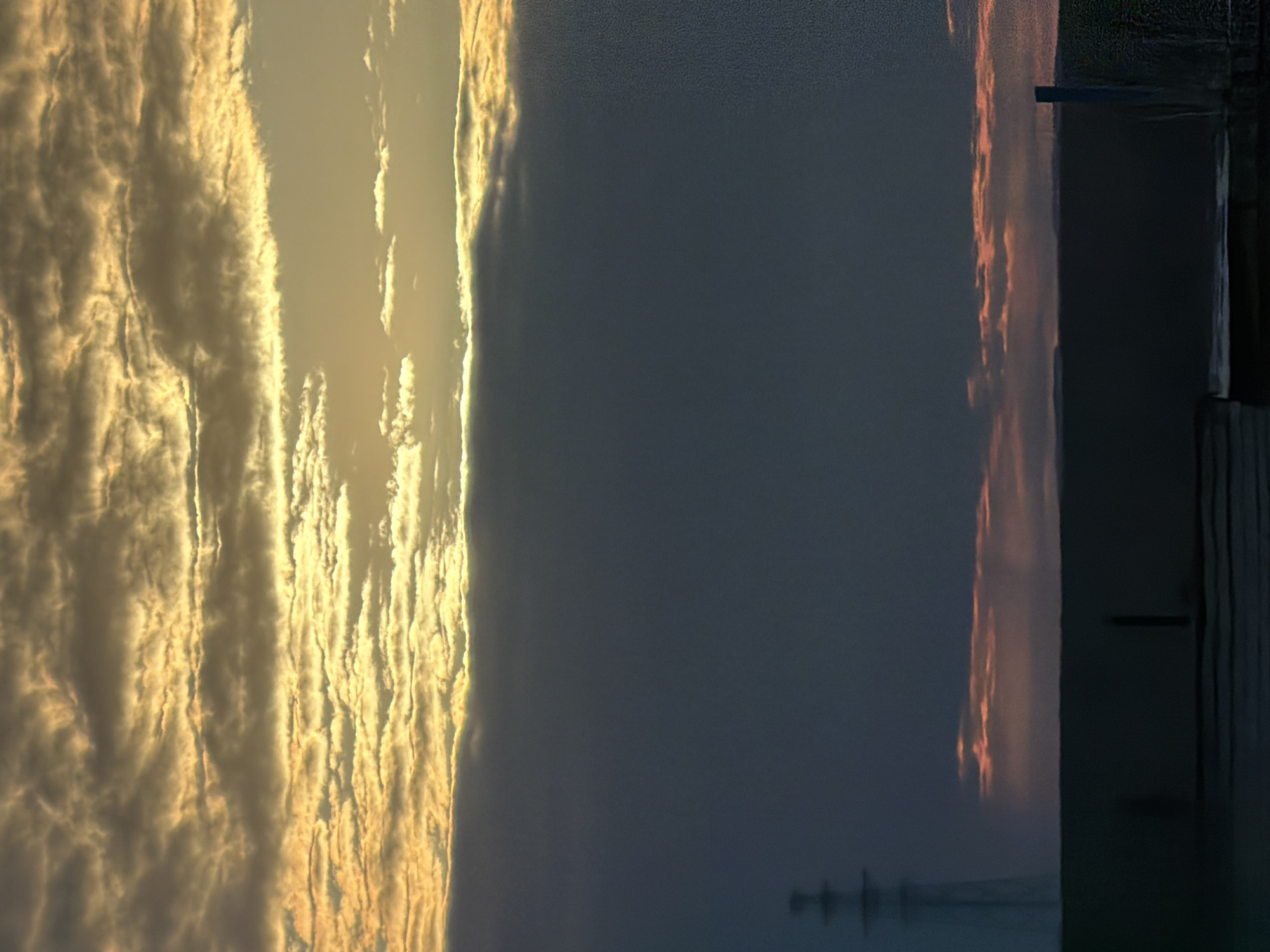 The image shows a sunset scene in what appears to be Chaikovsky, Russia, with a view towards the Kama River. The left side of the image is dominated by golden, cloud-filled skies from the setting sun, while the right side is a darker blue sky. The river occupies a small portion on the right, reflecting the colors of the sunset. A pier or wooden structure is visible on the edge of the river. There is a faint structure visible in the water near the bottom edge.