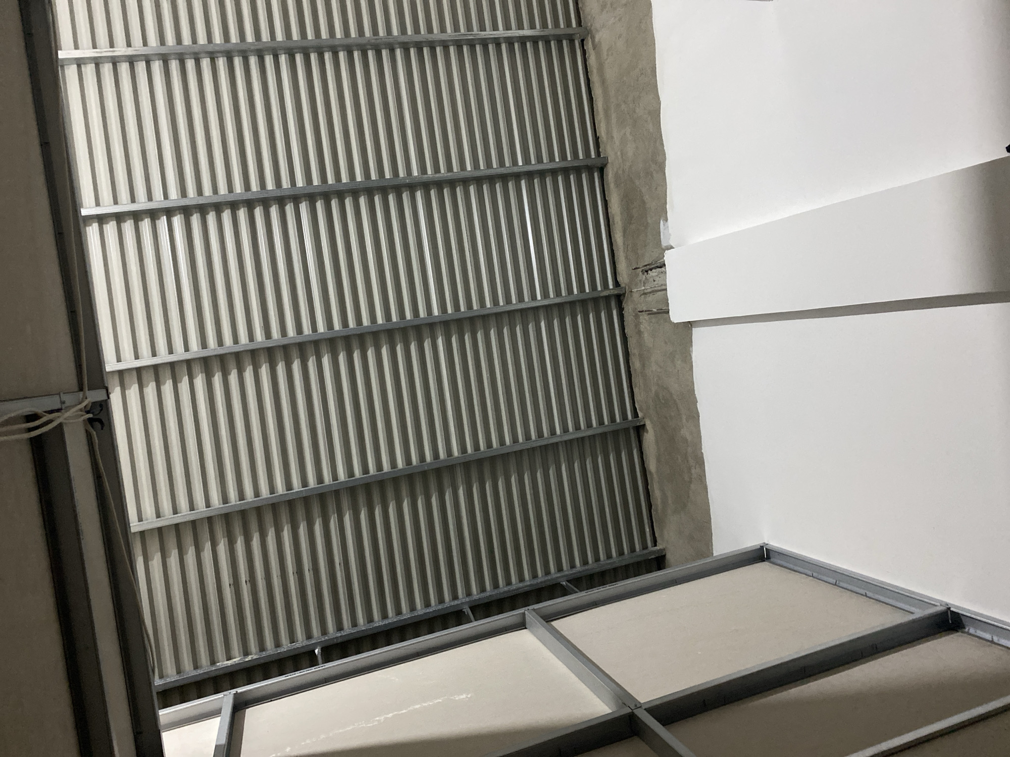 The image shows an interior view of an unfinished building. A corrugated metal roof is visible, supported by metal framing. In the foreground, a section of drywall is leaning against the metal framing. The floor appears to be concrete. The overall impression is of construction in progress. The location is identified as Ta', Indonesia. There are no people or specific actions depicted.
