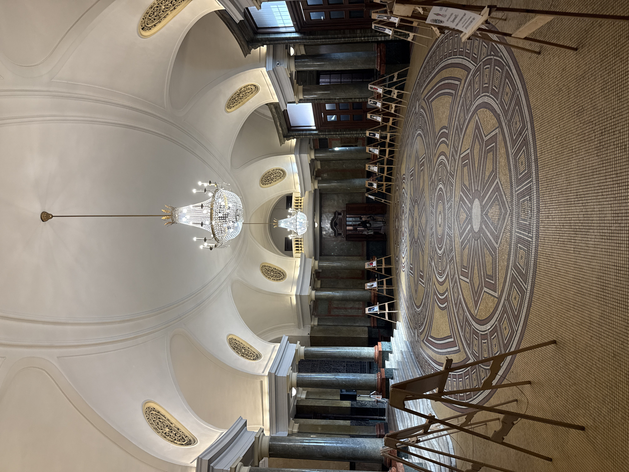 The image depicts the interior of a building in Sofia, Bulgaria. The architecture is grand and features a high, arched ceiling with ornate moldings and a central chandelier. Columns line the sides of the room, leading to a more dimly lit area in the background. The floor is covered in a patterned mosaic. Several wooden easels are positioned around the room, displaying photographs or documents.