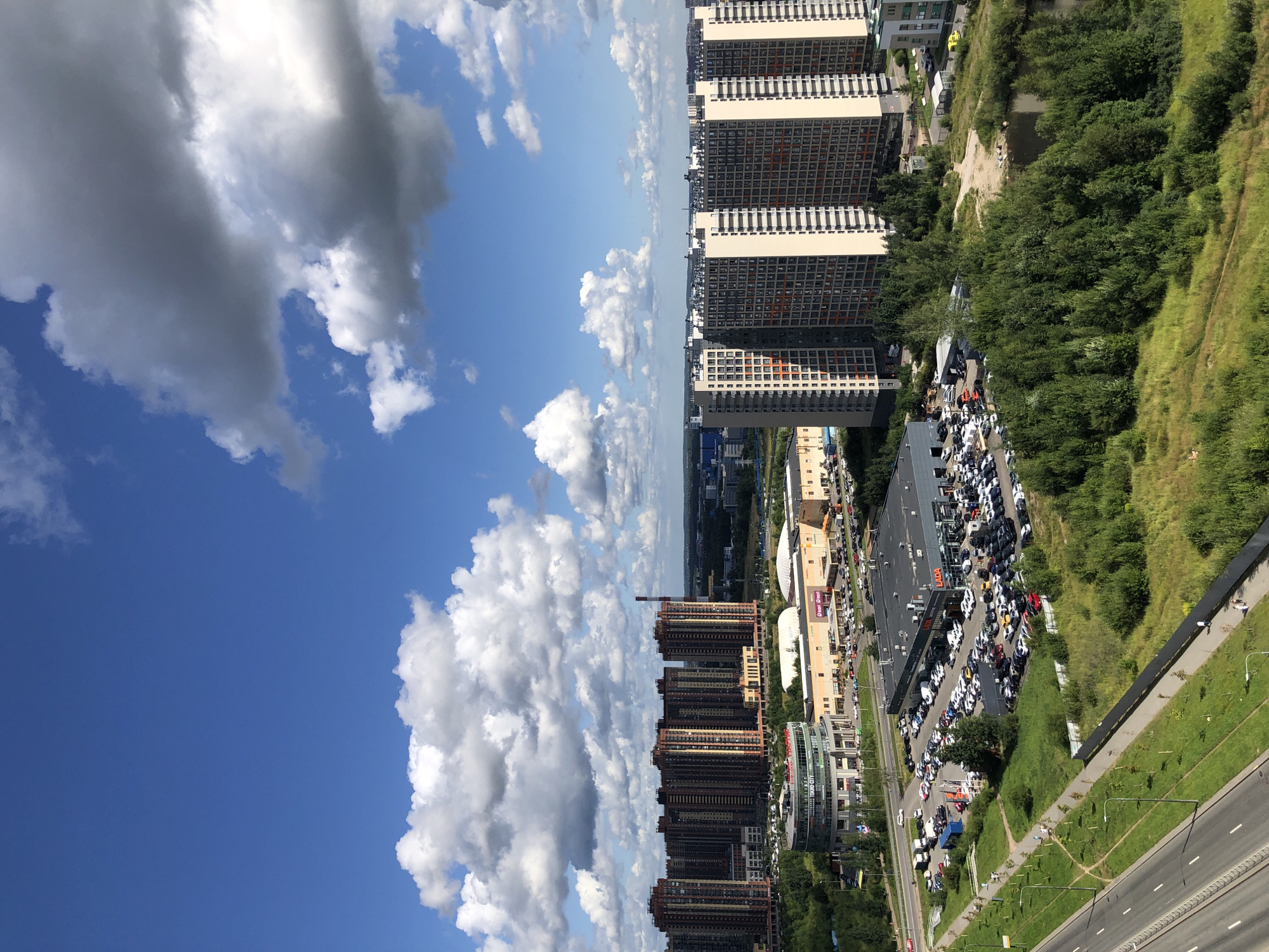 An aerial, full-shot, eye-level, wide-angle perspective captures a sprawling urban landscape under a bright, partly cloudy sky. The foreground features a multi-lane highway on the left, bordered by a grassy median and a sidewalk with lampposts. Adjacent to the highway is a large, open field of green grass and dense, tall green trees. Beyond the field, a parking lot is filled with numerous cars, situated in front of a low-rise, dark-roofed building with "LADA" signage visible. To the right of the parking lot is a small pond surrounded by lush green trees. In the middle ground, several commercial buildings are visible, including a circular building with a glass facade displaying "TOKKO-CITY" and "METRO" signage, as well as a longer, light-colored commercial building with a large, arched roof. In the background, a dense cluster of very tall, reddish-brown apartment buildings stands prominently. Further in the distance, more residential and industrial buildings are visible, stretching towards the horizon. The sky is a vibrant blue, dotted with fluffy white and gray clouds. No people are clearly discernible in the image, though distant figures may be present on the sidewalk. The overall scene depicts a modern, developing urban environment with a mix of residential, commercial, and infrastructural elements.