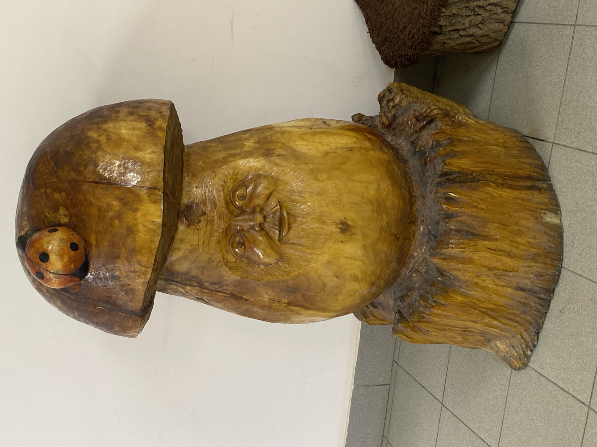 A large, intricately carved wooden sculpture is the central subject of the image. The sculpture depicts a human-like face emerging from what appears to be a mushroom or a natural wooden form. The face, carved into the lighter-colored wood, features closed or downcast eyes, a prominent nose, and a slight smile or neutral expression.
The "cap" of the mushroom form sits atop the head, made from the same polished wood, exhibiting visible wood grain and variations in golden-brown hues. A small, red and black painted ladybug figurine is attached to the top-left side of this cap, adding a whimsical detail. Below the face, the sculpture transitions into a rougher, darker wooden base, resembling tree roots or natural wood bark, providing a sturdy foundation.
The sculpture has a glossy, varnished finish that reflects light, highlighting the natural textures and lines of the wood. It is displayed indoors, against a plain white wall, and stands on a gray tiled floor with distinct grout lines. In the upper right background, another piece of wood or sculpture with a rough, bark-like texture is partially visible. No specific activity is depicted, and no text is visible in the image.