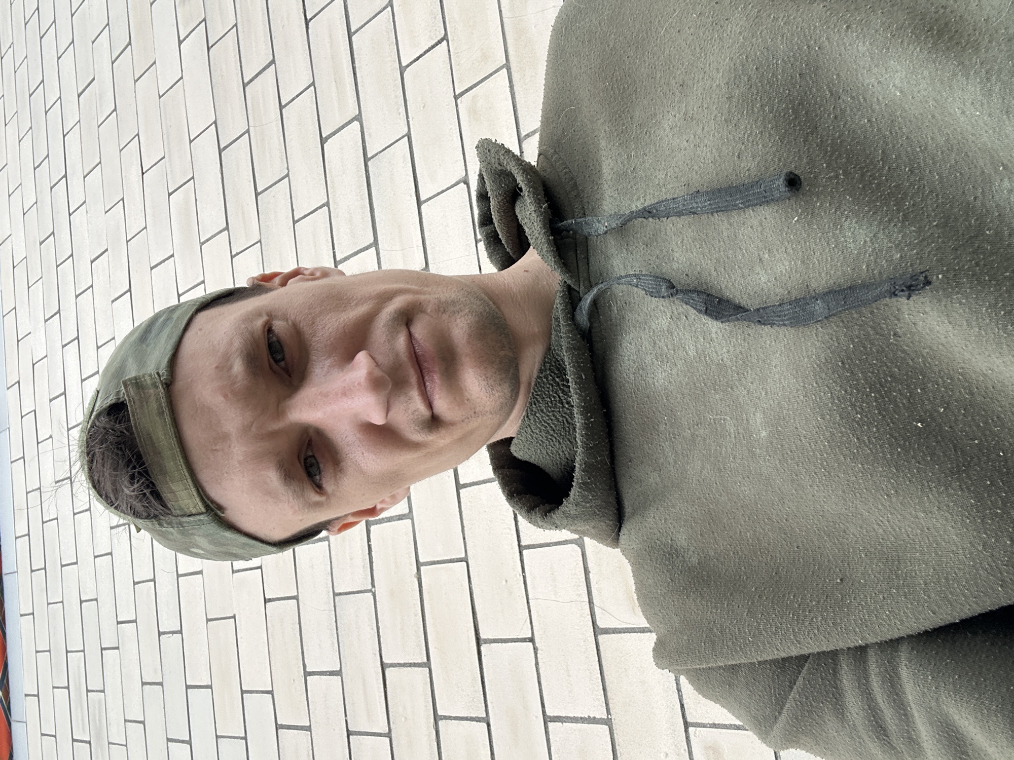 A close-up, high-angle selfie shows a Caucasian male wearing a green camouflage baseball cap and a faded olive green hooded sweatshirt. The man has short, light brown hair visible under the cap, light green eyes, and fair skin with some stubble. The background consists of a light-colored brick wall, with faint shadows suggesting an outdoor setting. The hoodie appears worn, with pilling on the fabric and frayed drawstrings. The image is shot from a slightly downward perspective. The exact location in Makiv, Ukraine, cannot be determined from the visual information provided.