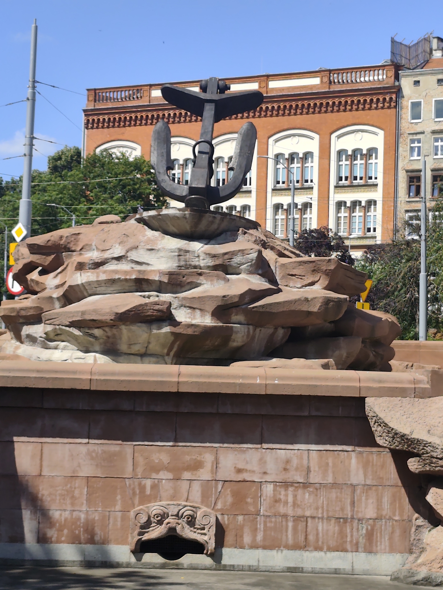 The image shows a fountain with an anchor sculpture on top. The fountain is made of large, layered stones that resemble a rock formation. Below the stone structure, there is a base made of reddish-brown bricks with a carved grotesque face acting as a water spout. In the background, there are buildings with orange and beige facades, some with white-trimmed windows. Trees and utility poles are also visible. The sky is clear and blue. This fountain is located in the city of Szczecin, Poland, specifically in the Solidarity Square (Plac Solidarności).
