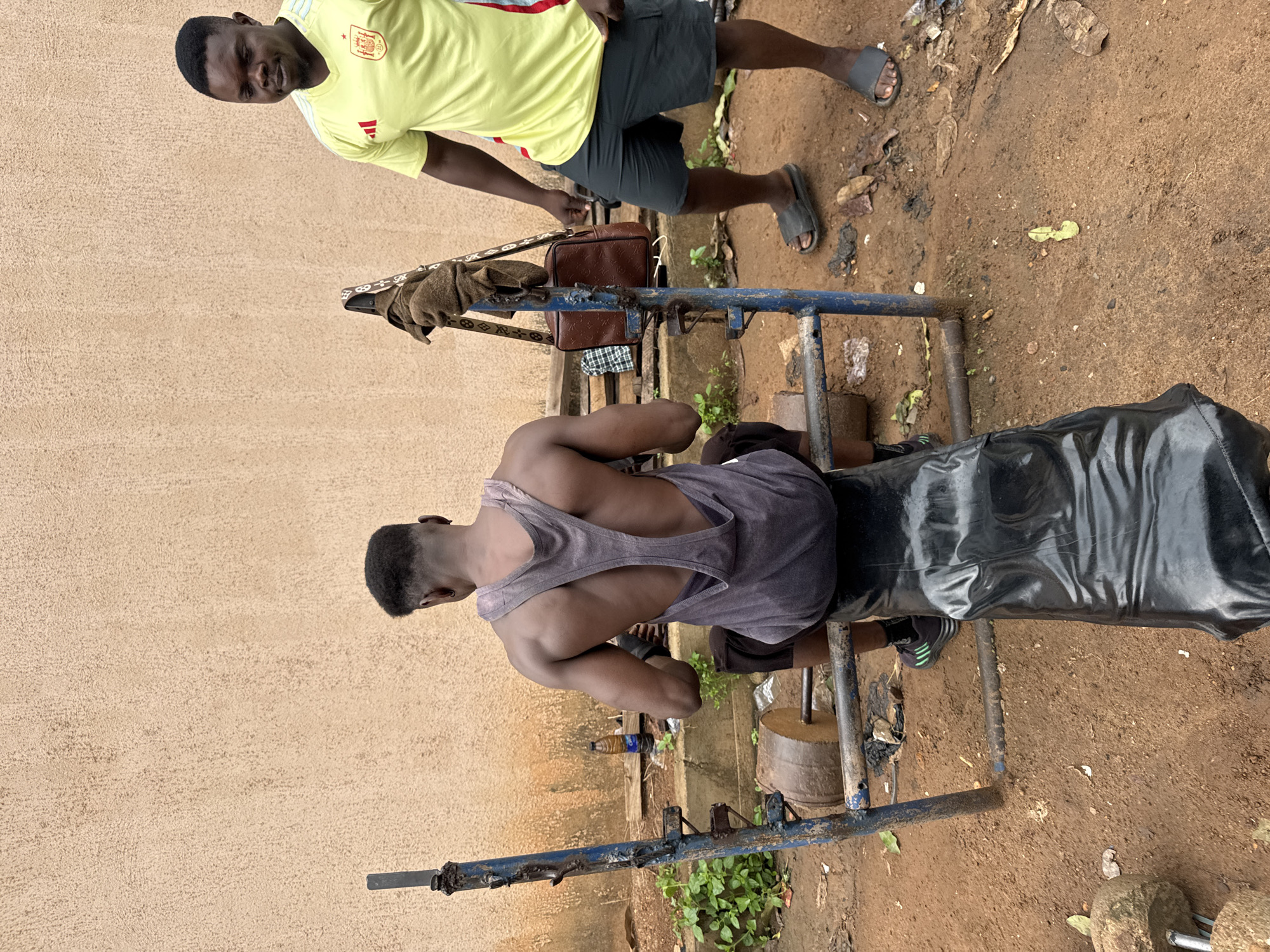 The image depicts an outdoor gym scene in Akwanga, Nigeria. Two men are present. In the foreground, a man with a muscular back is seated on a weight bench, wearing a grey tank top and black shorts. He appears to be mid-workout. To his right, a second man stands, wearing a bright yellow t-shirt with red accents and dark shorts. He is looking towards the left of the frame. The gym equipment is rudimentary, consisting of a metal bench and weight racks made from pipes and concrete. Weights made of concrete and metal are visible. The ground is dirt, and the background features a textured, tan-colored wall. Vegetation is growing around the gym equipment. A brown bag and a towel are hanging on a metal support.