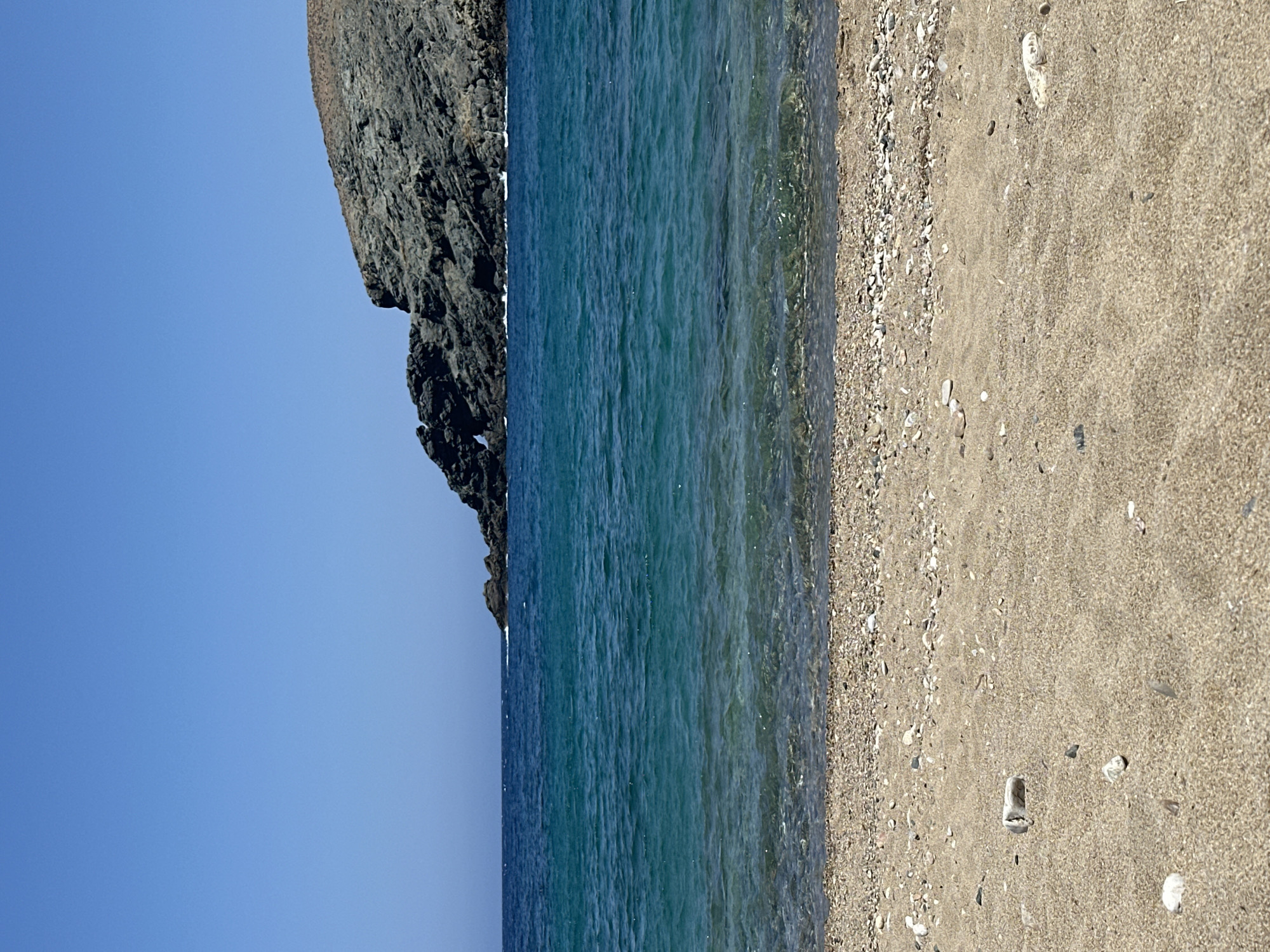 The image depicts a serene beach scene in Ano Mera, Greece, under a clear, bright blue sky. The foreground is dominated by a sandy beach with scattered small pebbles and stones. Beyond the sand, a line of larger pebbles marks the transition to the sea. The water is a vibrant turquoise, clear enough to reveal the seabed near the shore. Further out, the water deepens to a rich blue. In the background, a rugged, rocky cliff face juts out into the sea, with a distinctive archway carved into its side. The cliff is sparsely vegetated with dry, brown plants. There are no people or animals visible in the image. The overall impression is one of natural beauty and tranquility.