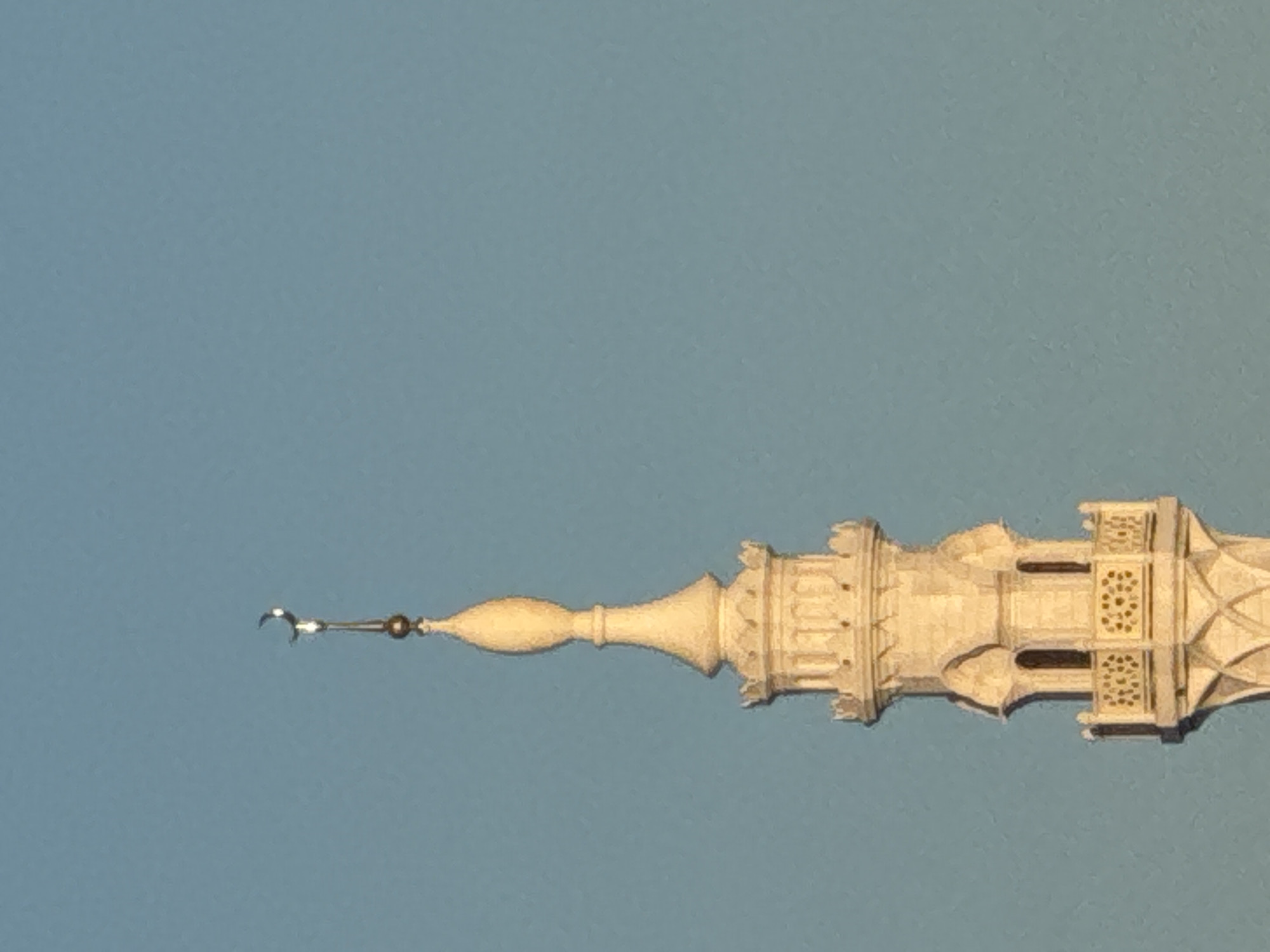 The image shows the top of a minaret, a tall, slender tower attached to a mosque, often found in Islamic countries. The minaret is made of white stone and is decorated with intricate carvings and architectural details. At the very top of the minaret, there is a crescent moon, a symbol of Islam. The minaret stands against a clear blue sky. This minaret is located in the city of Bolgar, Russia, which is known for its historical significance as an early Islamic center. The photograph captures the minaret in daylight, with the sun casting a warm glow on the stone. The focus is on the upper part of the minaret, with the sky occupying the majority of the frame.