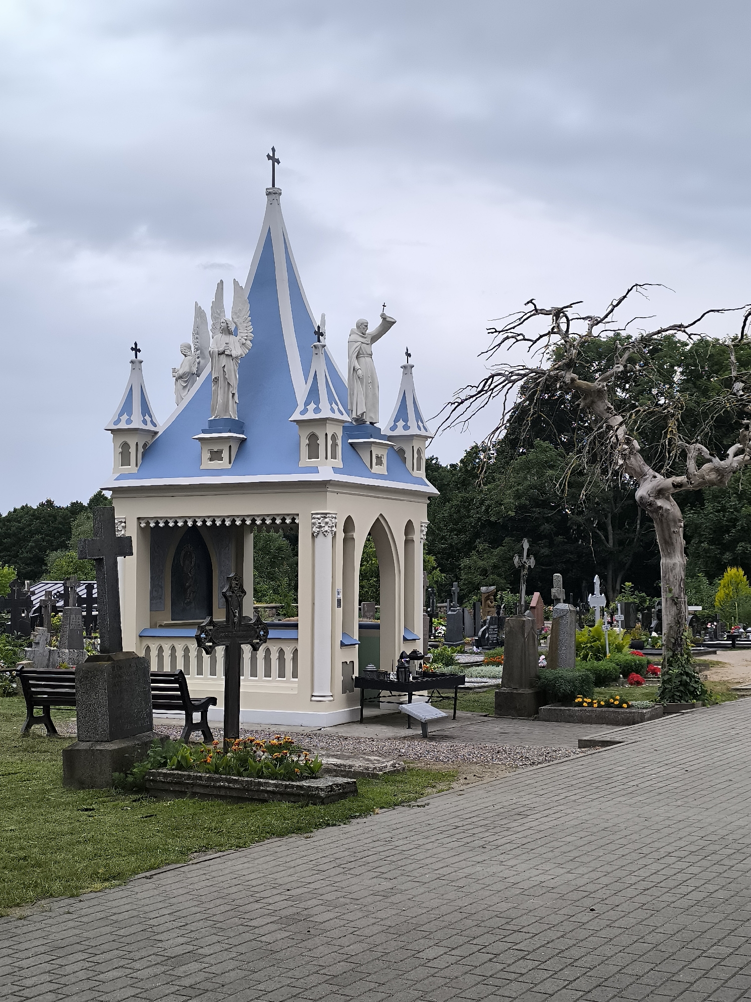 This image captures a section of the Kretinga cemetery in Kretinga, Lithuania. The focal point is a small, ornate chapel or mausoleum, painted white with a blue roof and adorned with statues of angels and figures. The chapel features a pointed roof with decorative spires and a cross at its apex.
Surrounding the chapel are numerous gravestones and monuments of various shapes and sizes, typical of a cemetery. Several of these are crosses, some simple and stone, others more ornate and dark metal. There are also benches placed near some of the graves.
In the foreground, a paved walkway made of grey interlocking bricks leads into the cemetery. To the left of the walkway, a large, dark stone cross stands prominently, accompanied by a dark bench. A smaller, dark metal cross is positioned in front of the chapel.
A distinctive feature of the scene is a gnarled, leafless tree to the right of the chapel, its bare branches creating a stark silhouette against the overcast sky. The ground is covered in grass and patches of soil, with some flowers blooming around certain graves. In the background, a dense line of green trees forms a natural boundary for the cemetery. The sky is overcast with a neutral grey hue.