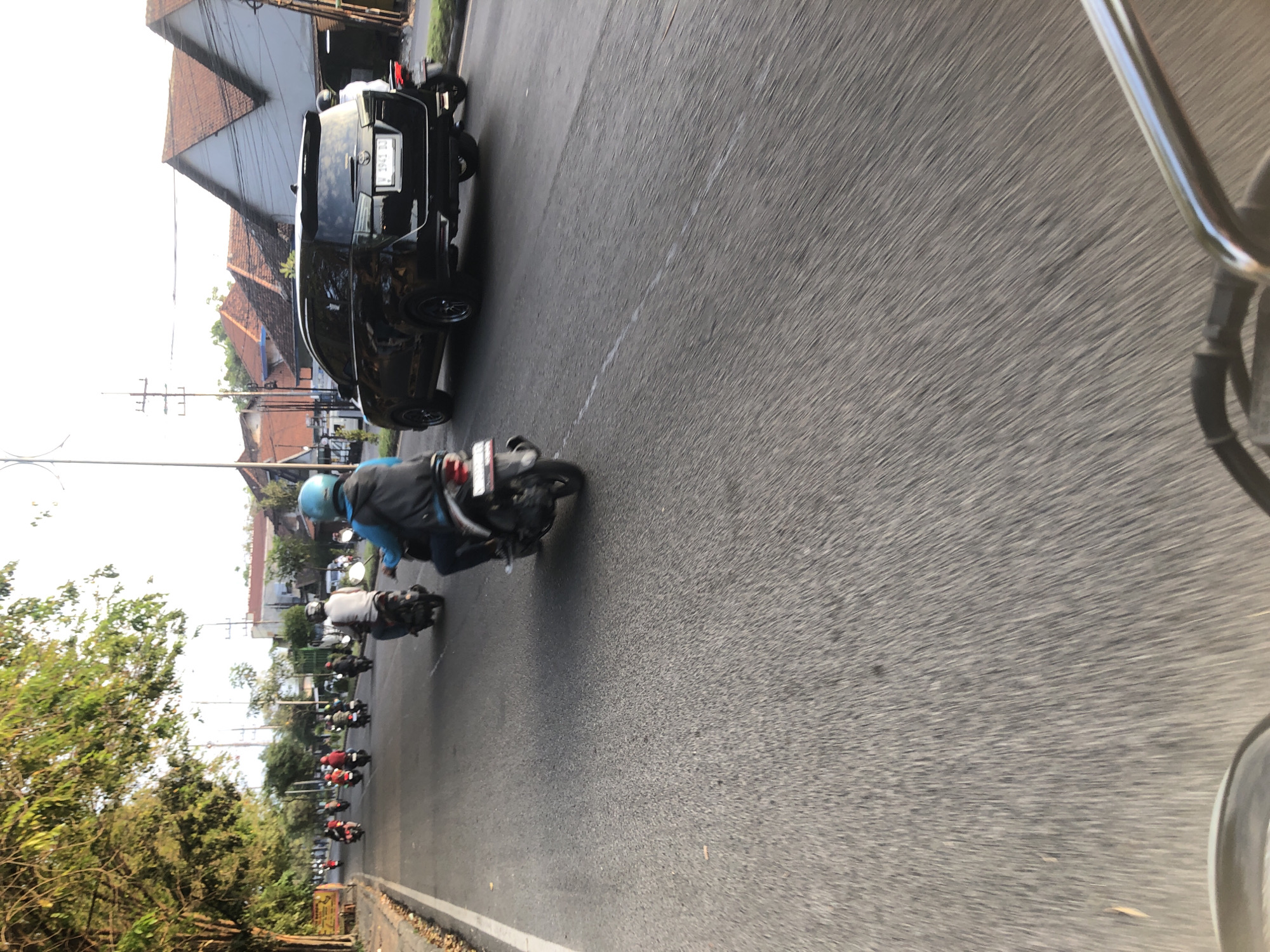 The image depicts a busy road in Kludan, Indonesia, with multiple motorbikes and a black SUV. The perspective is from a rider of a motorbike, looking forward. In the foreground, a portion of the motorbike's handlebars is visible, indicating movement. The road surface is asphalt with a white line marking the lane.
Several motorbikes are traveling in the same direction, with riders wearing helmets and casual clothing. One rider in the middle ground is wearing a blue helmet and a backpack. A black SUV is in the right lane, with its license plate partially visible. The background shows buildings with tiled roofs, trees lining the road, and utility poles. The sky is bright, suggesting it is daytime, possibly late afternoon due to the warm lighting. The overall scene conveys a sense of daily commute or travel on a typical Indonesian road.