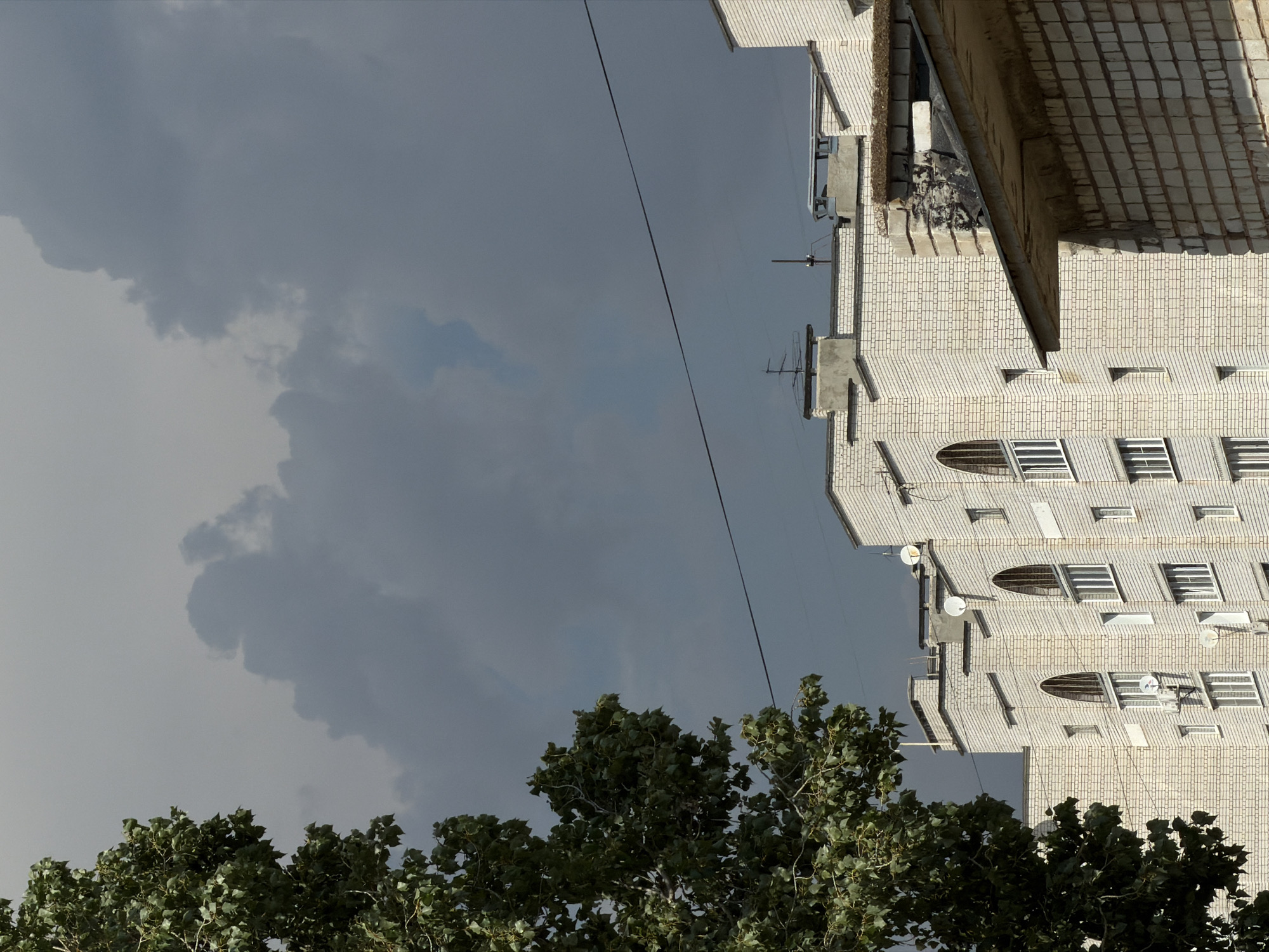 This is a medium-shot, eye-level, vertical photograph of a modern apartment building in Kamyshin, Russia, on a cloudy day. The building, constructed from light-colored bricks, features multiple stories with rows of windows. Some of the windows are arched, while others are rectangular. Several satellite dishes and antennas are visible on the rooftops. A large green tree with dense foliage is situated on the left side of the frame, partially obscuring the building. A thin black wire stretches horizontally across the upper portion of the image. The sky is filled with dark, dramatic clouds, suggesting an impending storm. The overall atmosphere is overcast and somewhat somber.