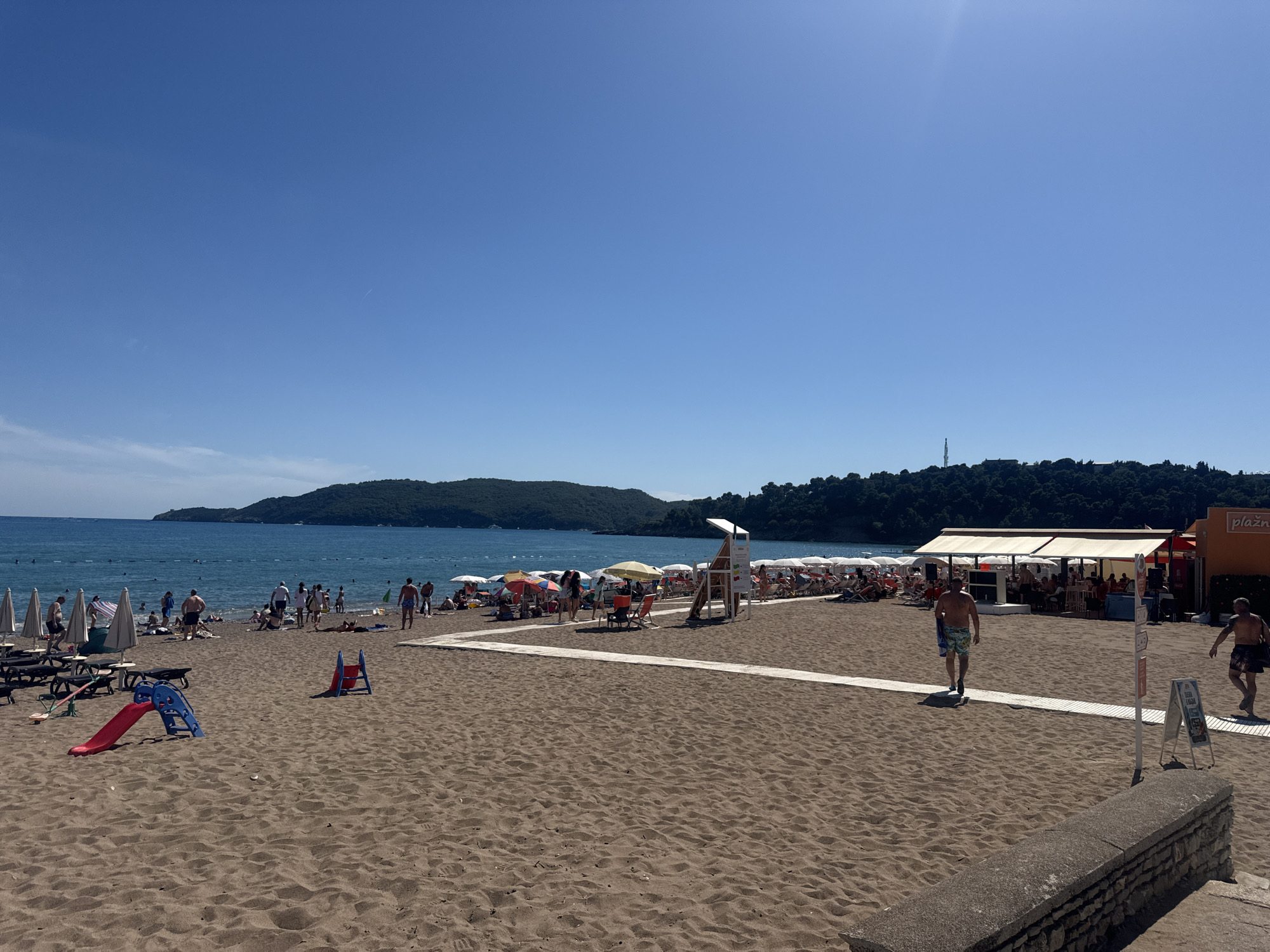 A wide shot of a sandy beach in Boreti, Montenegro. The beach is populated with beachgoers, some swimming or sunbathing, while others are gathered under umbrellas. There are several rows of beach chairs and umbrellas set up on the sand. A children's playground with a red and blue slide is visible in the foreground. The sea is a clear blue, with a distant forested hill on the horizon. A building with an orange facade and the word "PLAZA" visible is located on the right side of the image. The sky is clear and blue.
