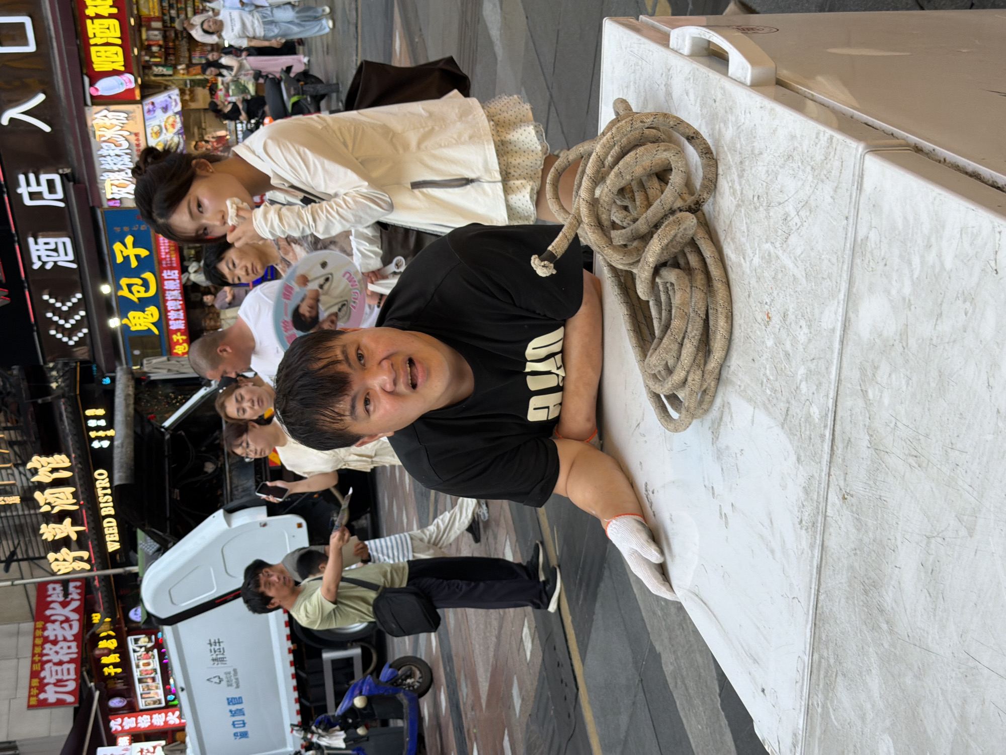 In Yuzhong District, China, a man wearing a black t-shirt and white gloves leans on a white surface, possibly a cooler or a refrigerator, with a thick coil of rope on top. His facial expression is one of surprise or exertion. Behind him, a young woman in a white jacket is eating something, while other people and businesses are visible in the background. A sanitation truck is parked on the street, suggesting a commercial or urban setting.