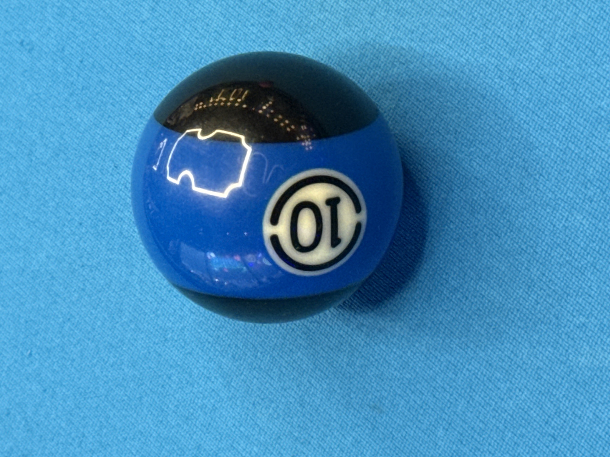 A blue and black pool ball, labeled with "10" in a circle, rests on a light blue surface. The ball casts a shadow to its right. The location is not discernible from the image.
