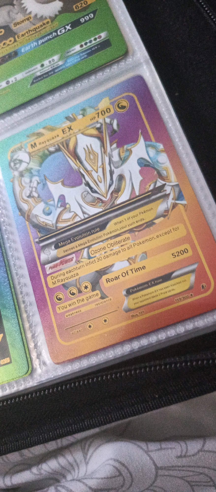 The image shows a close-up of two Pokemon cards inserted into a plastic binder. The card in the foreground, which is a Mega Rayquaza EX card, is predominantly purple and gold with an illustration of the Pokemon. Text on the card includes "HP 700", "Mega Evolution rule", "Ozone Obliterate", "Roar Of Time", and "Pokemon-EX Rule". The card in the background is green and has text such as "Stomp", "Earthquake", and "Earth punch GX". The binder appears to be black with a textured surface on the right side, and the cards are placed in transparent plastic sleeves. The background is out of focus but appears to be a dark surface with a patterned fabric underneath. There is no discernible connection to the city of Brwinów, Poland.