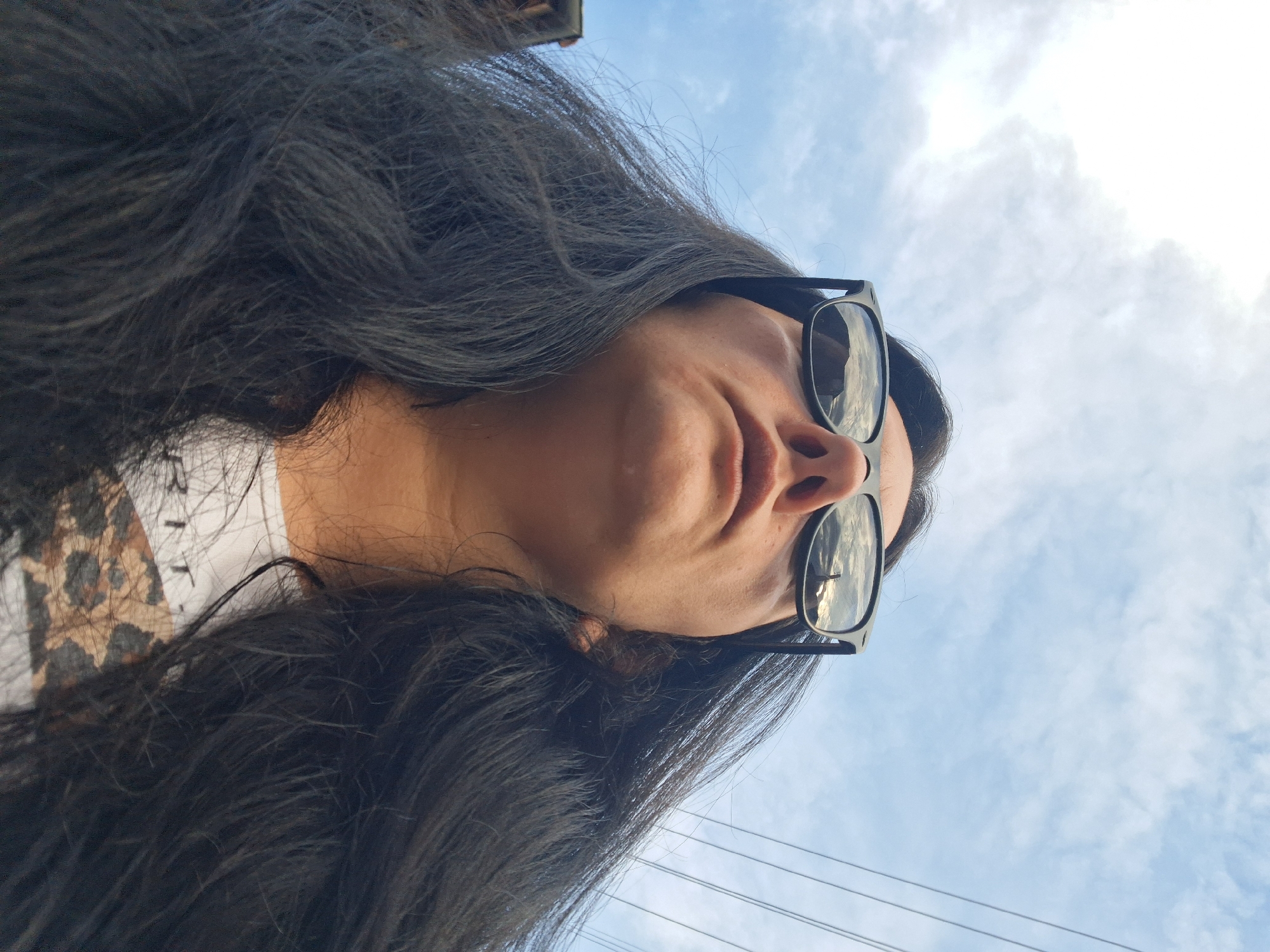 A low-angle, close-up, selfie-style photograph features a person wearing black-rimmed sunglasses, looking upwards towards a cloudy blue sky. The reflection in the sunglasses shows a bright sky with some clouds and what appears to be a tower or a tall structure. The person has long, dark, wavy hair that frames their face and flows down. Their mouth is slightly open, and they are wearing a white t-shirt with a leopard print design. The image captures the person from the chest up, with the sky dominating the background, interspersed with some overhead power lines. The scene is likely outdoors, with the low angle suggesting the photo was taken while lying down or standing very close to the ground. The location is not specified in the image itself.