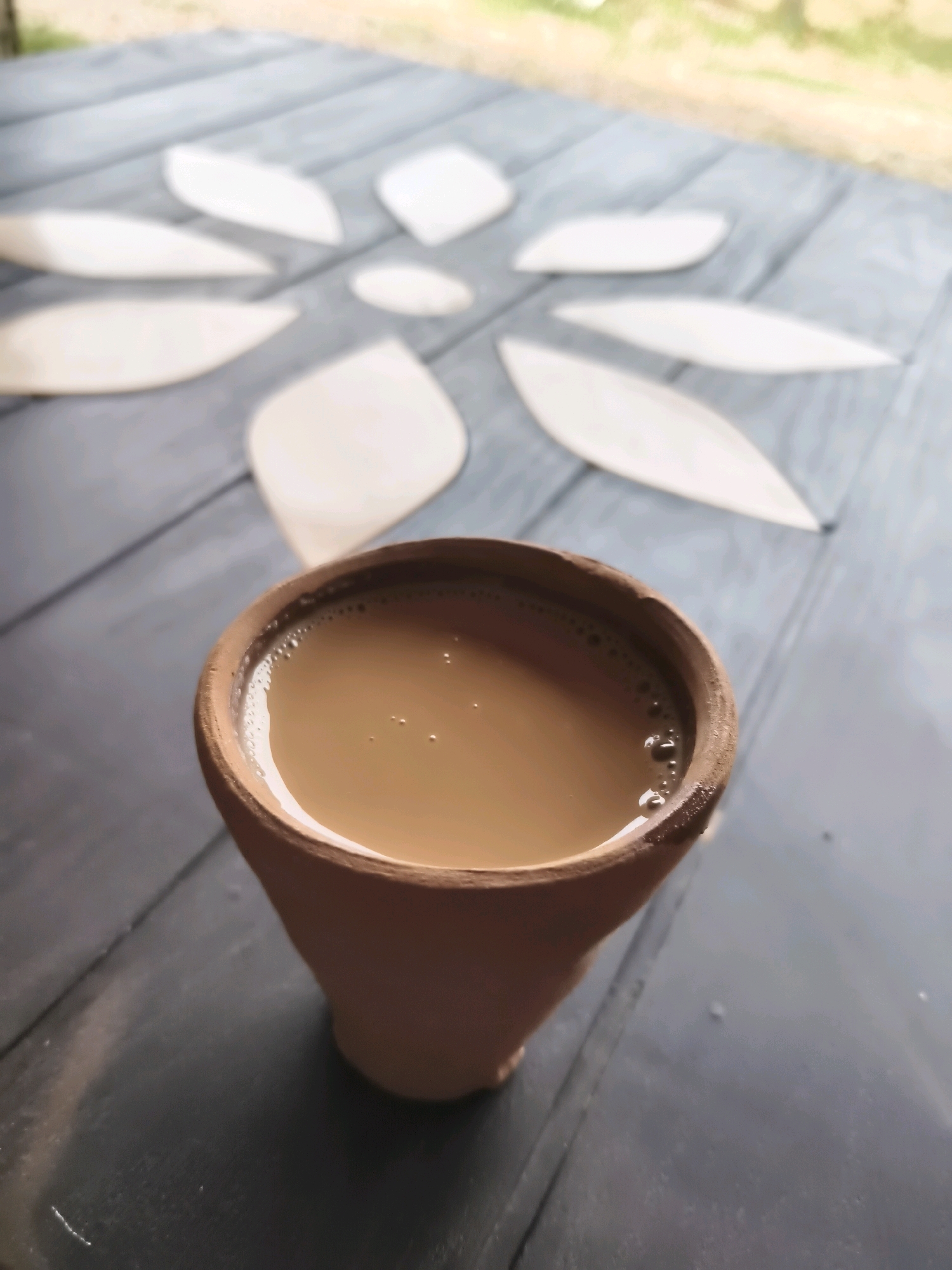 A close-up, high-angle shot shows a clay cup filled with tea sitting on a dark, wooden table. The tea has a light brown color and a foamy surface with small bubbles. The cup is made of unglazed clay and has a tapered shape. Behind the cup, on the table, there is a pattern of white, petal-shaped objects arranged in a circular, floral design. The table appears to be outdoors, with a blurred background suggesting greenery and natural light. The overall impression is one of a simple, tranquil setting.