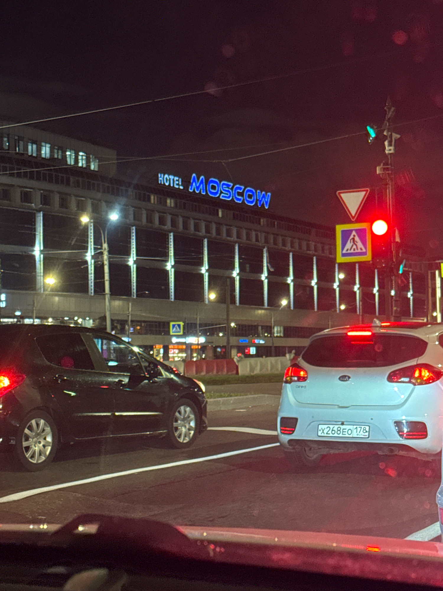 The image shows a night-time scene in Saint Petersburg, Russia, in front of the Hotel Moscow. In the foreground, the rear of a white Kia hatchback is visible, with its license plate reading "X268EO 178 RUS". To the left, a black car, possibly a hatchback, is also visible. Traffic lights are illuminated, with a red light prominently displayed. A pedestrian crossing sign is also visible. The Hotel Moscow building is lit up with blue neon signs and architectural lighting. The overall scene depicts a city street at night, likely experiencing traffic congestion due to the red traffic light.