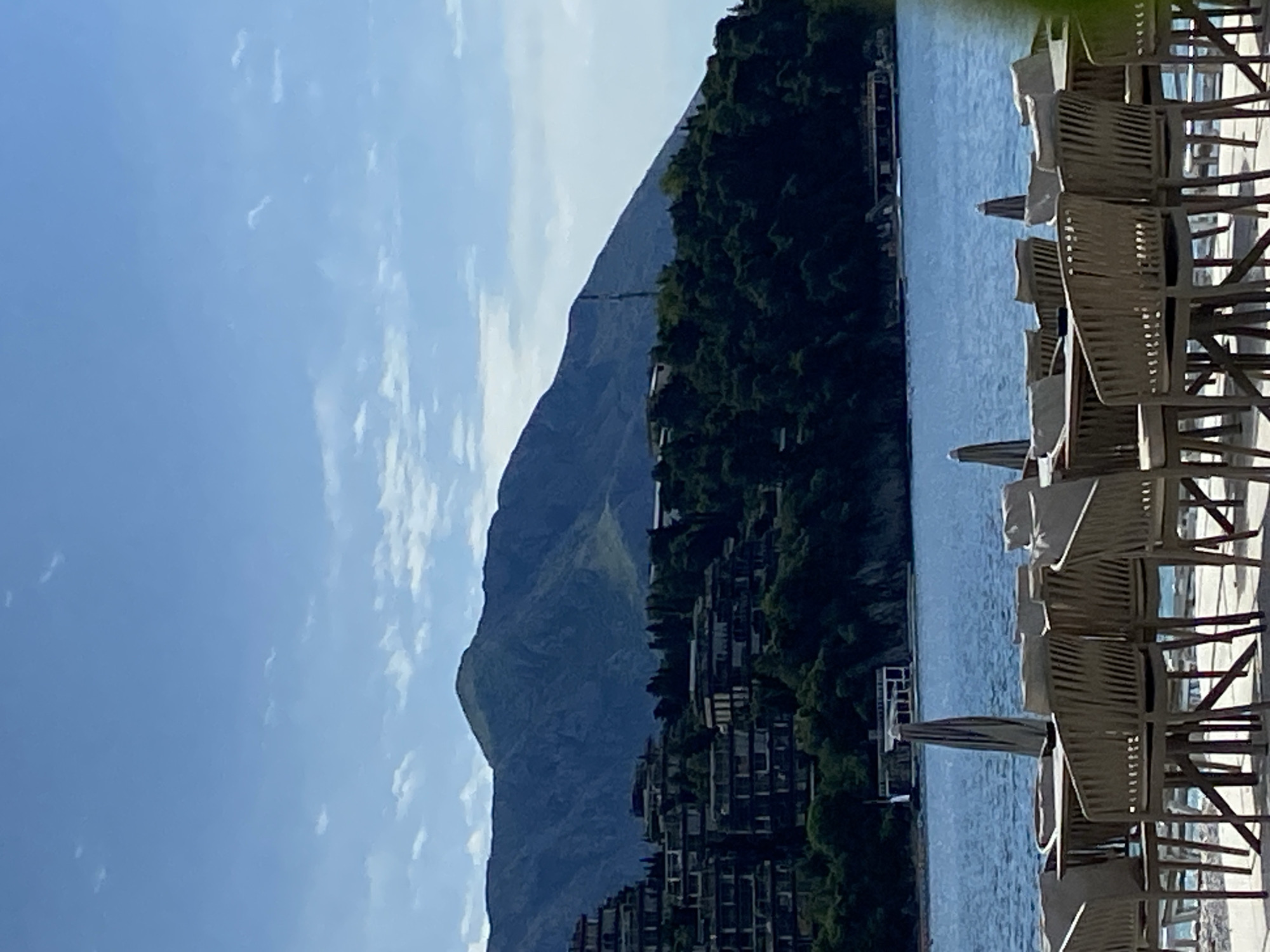A scenic view showcases a mountain backdrop, a lake, and beach chairs. The mountain, primarily in hues of gray and blue, contrasts with a green, tree-covered area. In the lake, there are a couple of small docks. A row of beach chairs, under white umbrellas, is located on the right side of the image. A portion of a blue sky with some clouds is visible above the mountain. The image appears to be taken in Budva, Montenegro, given the setting.