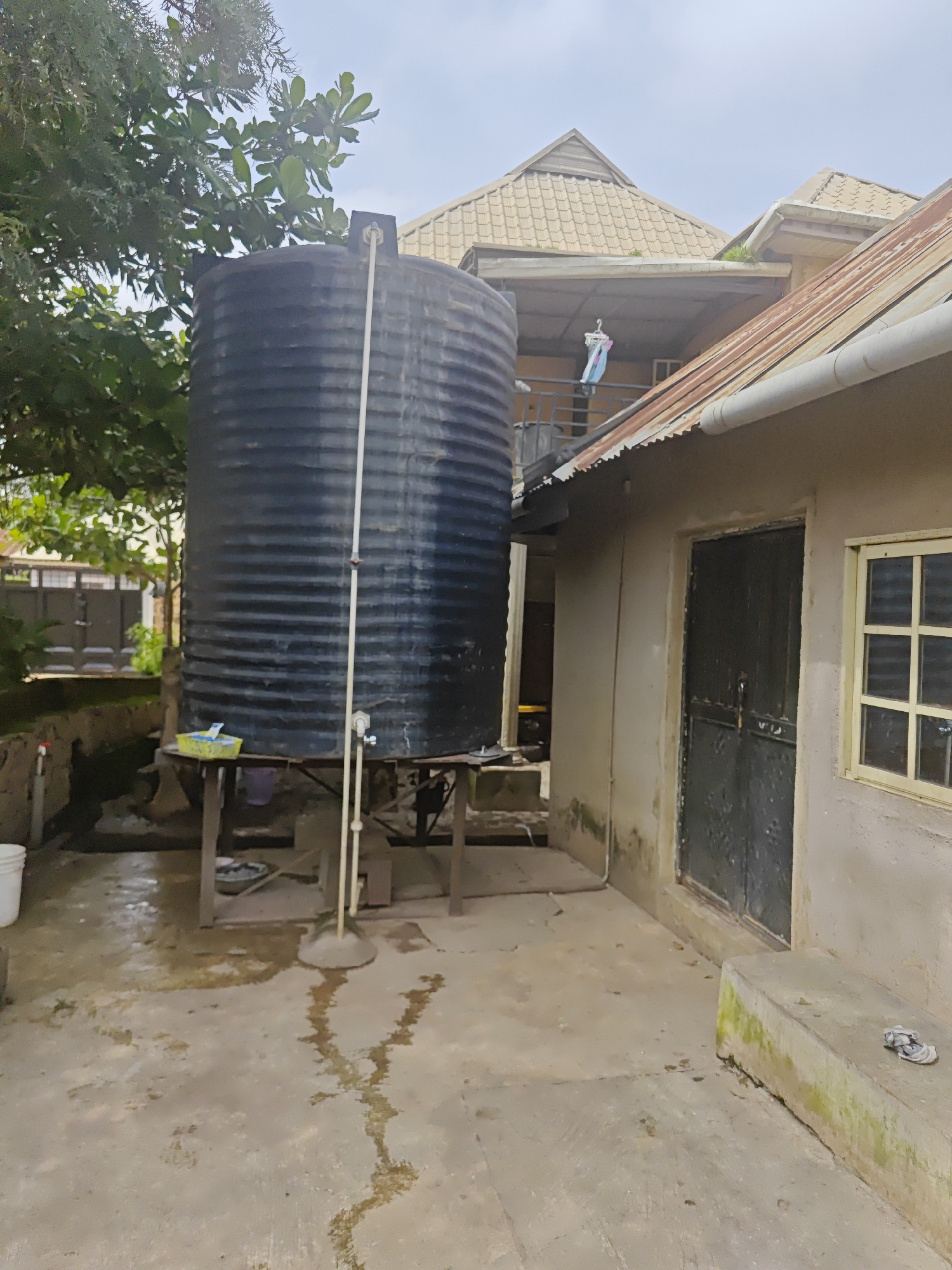 The image depicts an outdoor scene in a residential setting, likely in Bukuru, Nigeria. The primary subjects are a large, ribbed black water tank elevated on a rustic metal stand and an adjacent building. The tank is a prominent feature, suggesting a need for water storage. Below the tank, there's a small table holding a yellow plastic basket and a purple bucket, with a white bucket visible to the left of the frame. The ground is paved and appears wet, with water stains forming patterns on the concrete.
The building features a light-colored exterior wall, a dark metal double door, and a window with multiple panes. The roof of the building is made of corrugated metal, showing signs of rust. Behind the water tank, another building with a patterned, yellowish roof is visible, with a clothesline with colorful laundry hanging from it. Green foliage from trees partially obscures the upper portion of the water tank and the sky.
The lighting suggests it's daytime, possibly overcast or after rain, given the wet ground. There are no people visible in the image. The scene appears to be a functional, perhaps utilitarian, area of a home or compound, focusing on water supply and basic housing structures. The overall impression is one of a pragmatic living environment where water management is important.