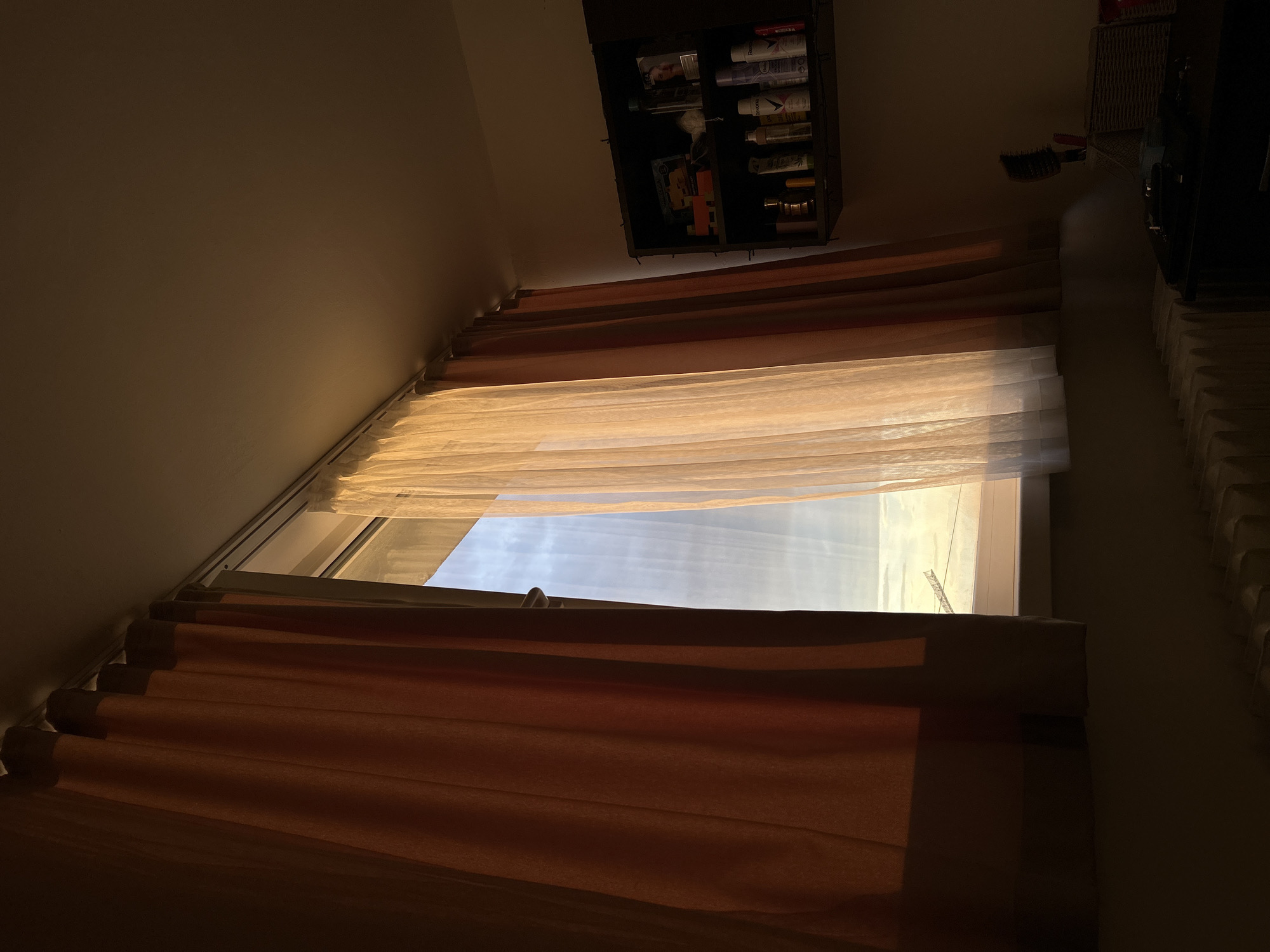 The image, presented with a 90-degree clockwise rotation, depicts a dimly lit indoor scene focused on a window. Natural light streams in from the outside, illuminating a sheer, light-colored inner curtain that covers the upper portion of the window. Below this, a thicker, brownish-orange outer blind or curtain is partially drawn, covering the lower half of the window.
Through the window, beyond the sheer curtain, a bright, possibly cloudy sky is visible. On the far right, a section of what appears to be a construction crane is faintly discernible against the bright background.
Above the window (which would be on the ceiling if the image were correctly oriented), a dark, open cabinet or shelf is mounted. It holds various bottles and containers, with "Razo" faintly legible on one of the items. To the right of the window (on the right wall in correct orientation), a dark hairbrush hangs, and further right, a portion of a white radiator or similar structure is visible.
The room itself is cast in deep shadows, with the window serving as the primary light source. The warm quality of the light suggests it is daytime, possibly morning or late afternoon. The scene conveys a quiet and still atmosphere in what seems to be a residential or private room in Sofia, Bulgaria. No people are present, and no specific activity is occurring.