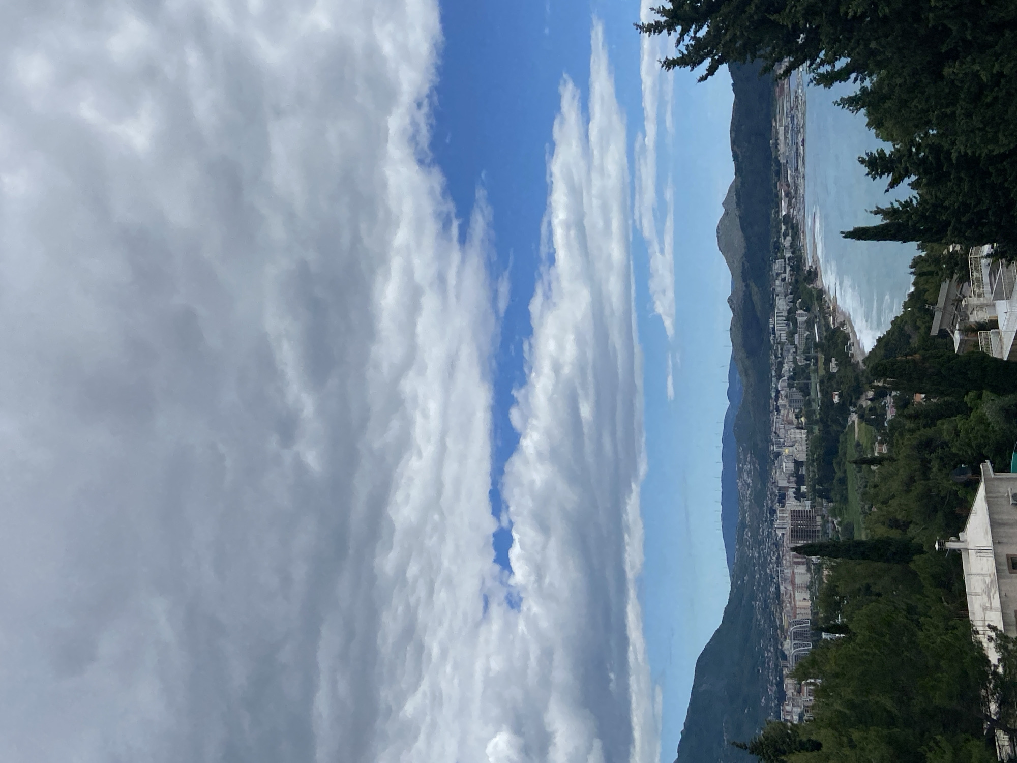 The image depicts a view from an elevated location in or near Šušanj, Montenegro, overlooking a coastal city. The foreground consists of greenery including trees and glimpses of buildings. A body of water, likely the Adriatic Sea, stretches along the shoreline, meeting a coastline dotted with buildings. Mountains are visible in the midground. The sky is filled with clouds, ranging from thin, wispy formations to denser, darker patches. The composition is oriented vertically.
