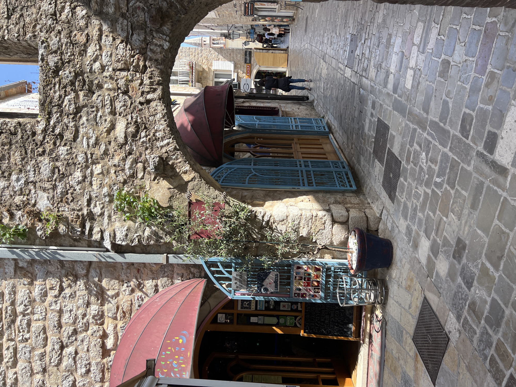 The image depicts a sunlit cobblestone alleyway in an old town, likely Budva, Montenegro. The perspective is from beneath a rough-hewn stone archway, looking down the pedestrian street. Buildings on both sides are constructed from rugged, light-colored stone, some with small plants growing from crevices.
On the left side of the alley, a building features a distinctive red, scalloped awning, with a second, similar awning closer to the camera displaying colorful painted dots. Below these, decorative blue metal gates or shutters are partially open, leading into establishments. A potted plant with an olive-like tree stands next to blue metal railings. Visible signs on these railings include "RESTORAN" and "LAVAZZA," along with small symbols indicating amenities like Wi-Fi and review platform ratings. A chalkboard menu lists food items such as "3x Kofus," "S.Klabu," "Pizza (2pcs)," "Fish klub," "Schrimps," "Birsteak," "Burger," and "Grill."
The alley floor is paved with irregular grey and brown cobblestones, interspersed with several dark metal grates. In the mid-ground, a person dressed in a dark jacket and jeans walks away from the viewer. Further down the street, additional individuals are visible, some seated at what appears to be outdoor dining tables, suggesting active businesses. Another shop sign further down reads "SHOP." The bright daylight casts distinct shadows, indicating a clear, sunny day. The scene portrays a vibrant, historic setting with pedestrian activity.