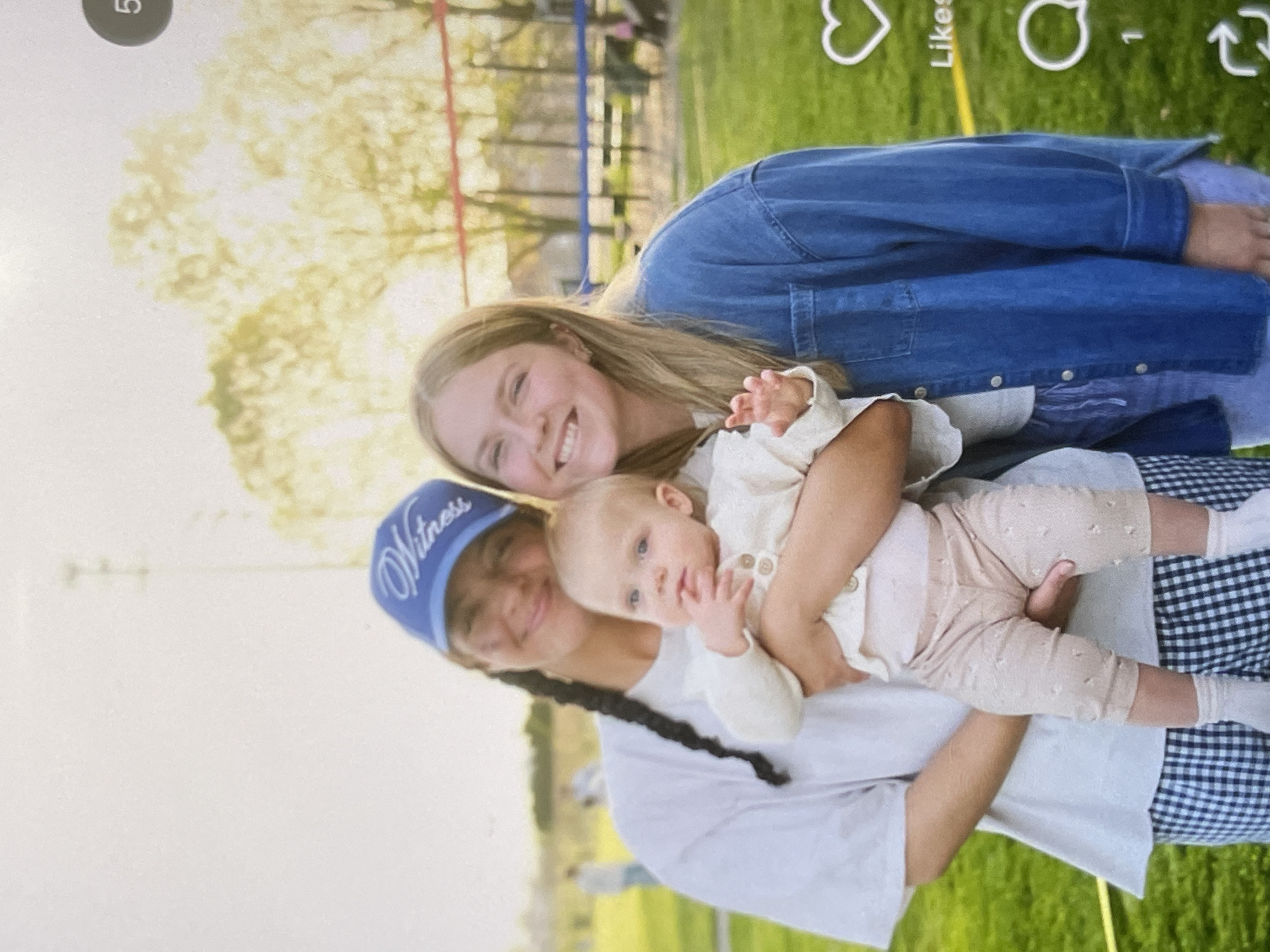 A medium shot captures two young women and a baby outdoors on a sunny day, with a blurred background suggesting a park or sports field. The woman on the left, wearing a white t-shirt and a blue "Witness" baseball cap, holds a baby dressed in a light-colored outfit. The baby is looking towards the camera with its right hand near its mouth. To the right, another young woman, wearing a denim jacket over a light-colored shirt, smiles at the camera. She is positioned behind and to the side of the baby. Both women are smiling and appear to be interacting with the baby. The ground is covered in green grass, and in the background, there are trees, a fence, and some distant structures. The lighting suggests it is late afternoon. A heart icon and the word "Likes" are visible on the right side of the image, indicating it is a screenshot from a social media platform.