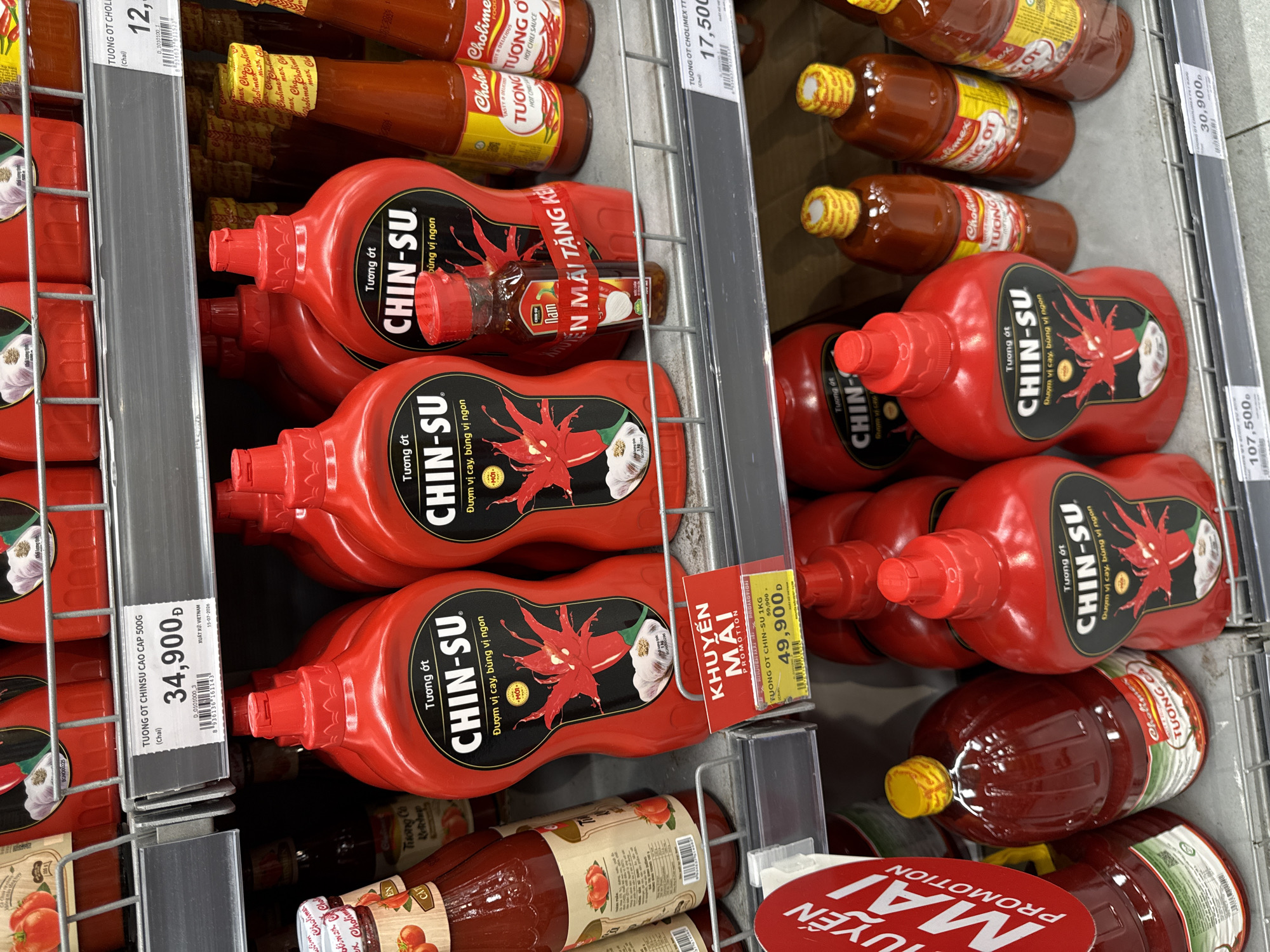 The image shows a shelf display of various hot sauces in a supermarket in Nha Trang, Vietnam. The dominant products are red bottles of "CHIN-SU" chili sauce. There are also bottles of "Cholimes" chili sauce visible. Price tags are displayed on the shelf, indicating prices in Vietnamese Dong. One tag reads "34,900₫" for a 500g bottle of "TUONG OT CHINSU CAO CAP". Another tag indicates a promotion for "TUONG OT CHIN-SU 1KG" at "49,900₫". The overall environment is a retail setting with metal shelving and fluorescent lighting. No people are visible in the image.