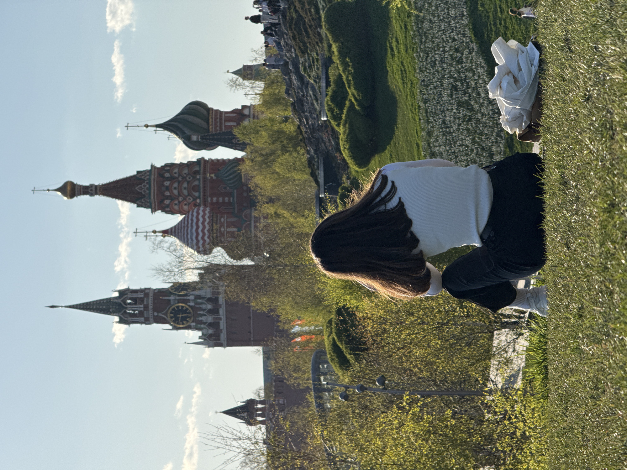 A young woman with long brown hair is crouching on a grassy area. She is wearing a white long-sleeved shirt and dark pants. A crumpled white garment is nearby. The background reveals the Moscow Kremlin and Red Square. The scene is set on a sunny day with a blue sky. There are trees in the scene.