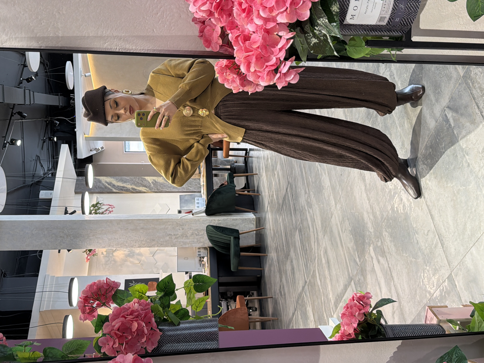 A woman is captured in a mirror selfie, posing in a modern interior space, likely a showroom or boutique in Krasnoyarsk, Russia. She is wearing a mustard yellow cardigan with prominent gold buttons, dark brown pleated wide-leg pants, and dark brown shoes. Her accessories include a dark brown beret, large gold hoop earrings, and rings on her fingers, as she holds a green smartphone to take the picture.
The background, reflected in the mirror, features light grey, marble-patterned floor tiles and walls. The ceiling is dark, possibly black, with several circular white lights and spotlights, suggesting contemporary indoor lighting. Dark green upholstered chairs with wooden legs are visible, along with glimpses of other furniture. The foreground of the mirror is adorned with lush clusters of artificial pink hydrangeas and green foliage in a patterned container. Visible text on display items includes "MODERN," "Acrylic Paints," "Fine Art Materials & Crafts," "CASHMERE," and a partial view of "FABRIC." The scene depicts a chic and well-appointed indoor environment.