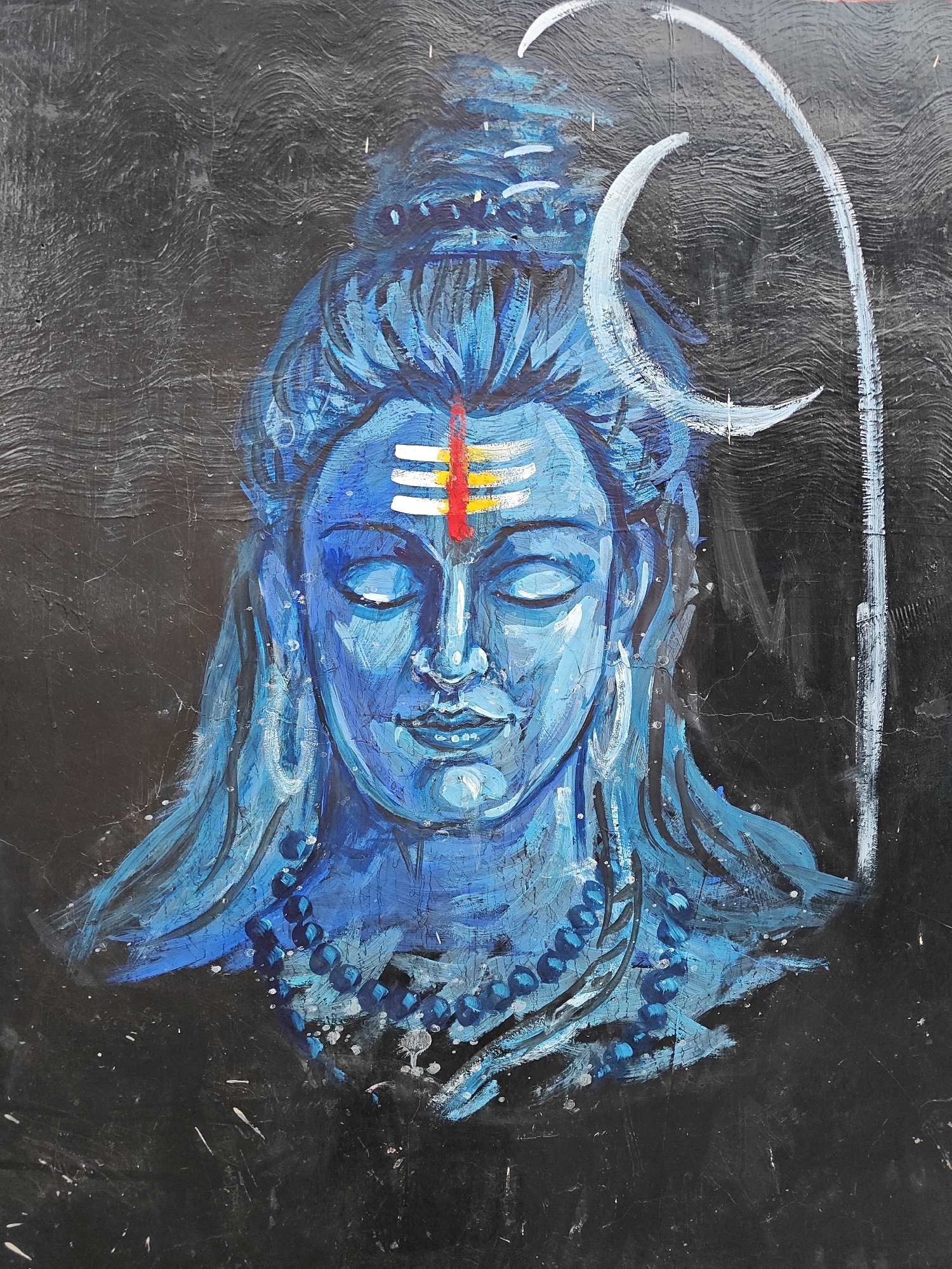 The image shows a painting of the Hindu deity Shiva, rendered in shades of blue against a dark, textured background. Shiva is depicted with closed eyes and a serene expression, wearing a beaded necklace and earrings. A crescent moon adorns his head, with a single white line extending upwards from it. His forehead is marked with a tilak, consisting of horizontal white lines and a red and yellow stripe. The style is somewhat rough and expressive, with visible brushstrokes. Based on previous analysis, it is highly probable this photograph was taken in Mumbai, India.