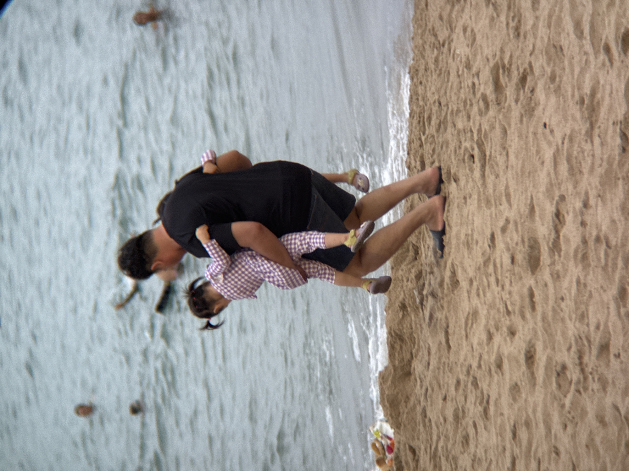 An adult male, wearing a black t-shirt and denim shorts, carries a young child on his back near the water's edge on a sandy beach. The child, wearing a pink and white checkered jumpsuit, is being held by the adult's arms. Both individuals are facing away from the camera, towards the ocean. The ocean in the background has moderate waves and several other people are visible in the water, some appearing to be swimming or wading. A small pile of items, possibly a bag and other beach accessories, lies on the sand to the left of the main subjects. The beach is composed of light brown sand with some uneven textures. The overall scene depicts a casual day at the beach. This scene is consistent with the beaches found in Nha Trang, Vietnam, known for its popular coastline and tourist activities.