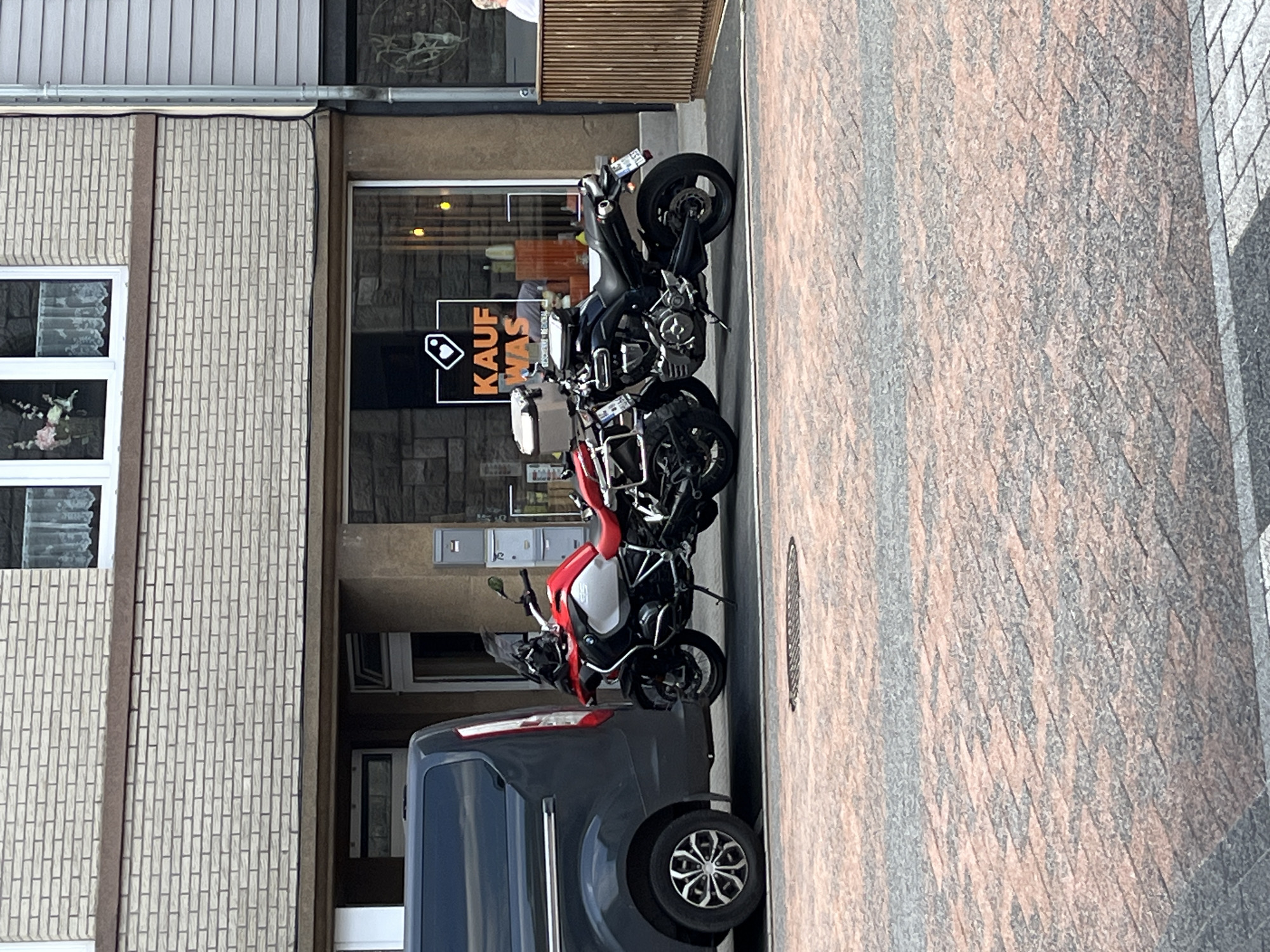 Two motorcycles are parked on a paved street in front of a building. The motorcycle on the left is a red and silver BMW GS adventure motorcycle. The motorcycle on the right is a black sportbike. A dark gray van is parked to the left of the motorcycles. The building behind the motorcycles has a brick facade with a large window displaying a sign that reads "KAUF WAS." The street is paved with reddish-brown and gray interlocking bricks. In the background, a person is visible standing at a counter inside the store. The scene appears to be in a commercial district.