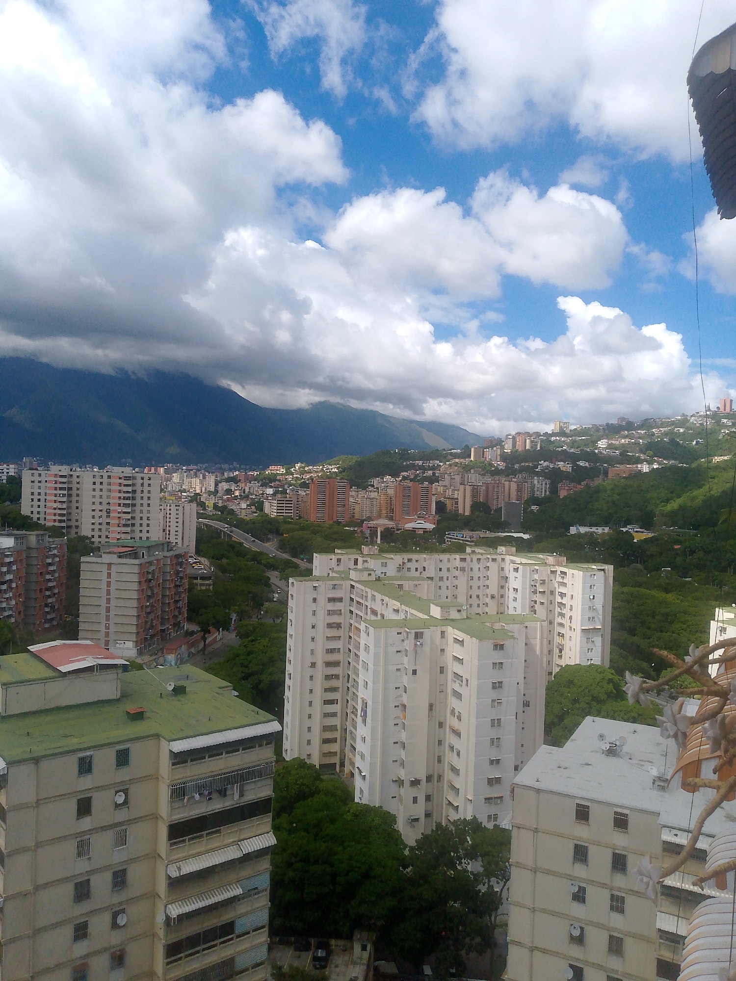 The image shows a panoramic view of a densely populated urban area in Caracas, Venezuela, with numerous high-rise apartment buildings and mountains in the background. The sky is partly cloudy with blue patches visible. The buildings are predominantly light-colored, with some featuring balconies and windows. Green trees are interspersed throughout the cityscape, especially in the foreground and on the hillsides. The overall scene suggests a typical residential neighborhood in Caracas, likely in an area with hilly terrain and close proximity to natural landscapes. No people or specific activities are discernible in this aerial perspective.