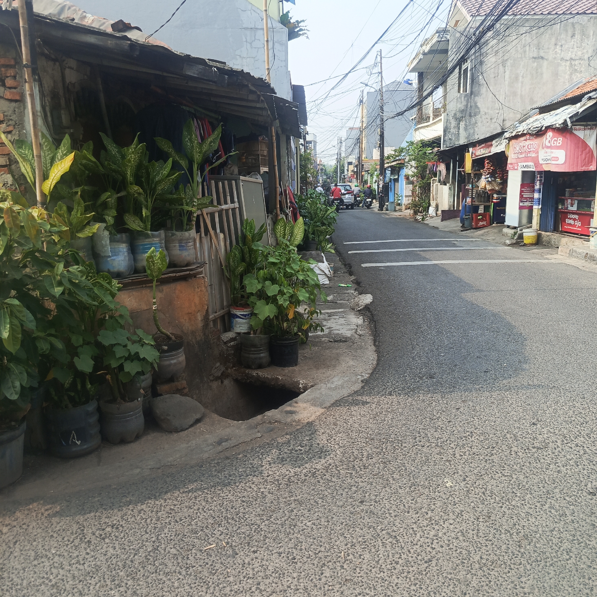 The image depicts a street scene in South Jakarta, Indonesia, likely a residential or commercial area with narrow roads and sidewalks. On the left, a rustic structure with a corrugated metal roof and exposed brickwork is visible, fronted by numerous potted plants, including Dieffenbachia and other leafy varieties. Some pots are made from repurposed plastic bottles. A drainage grate is present on the sidewalk in front of this structure.
The street itself is paved with asphalt and marked with white lines. Several motorbikes and a car are present on the road, indicating active local traffic. Buildings line the opposite side of the street, featuring a mix of modern and older architecture. Some of these buildings house small shops, one prominently displaying a red and white sign that reads "Telkomsel" and advertises "8GB" mobile data. Other shops show goods displayed outside, such as drinks in crates and packaged snacks. Utility poles with a dense network of electrical wires crisscross the sky, a common sight in urban areas of Indonesia. The overall impression is one of a vibrant, lived-in urban environment.