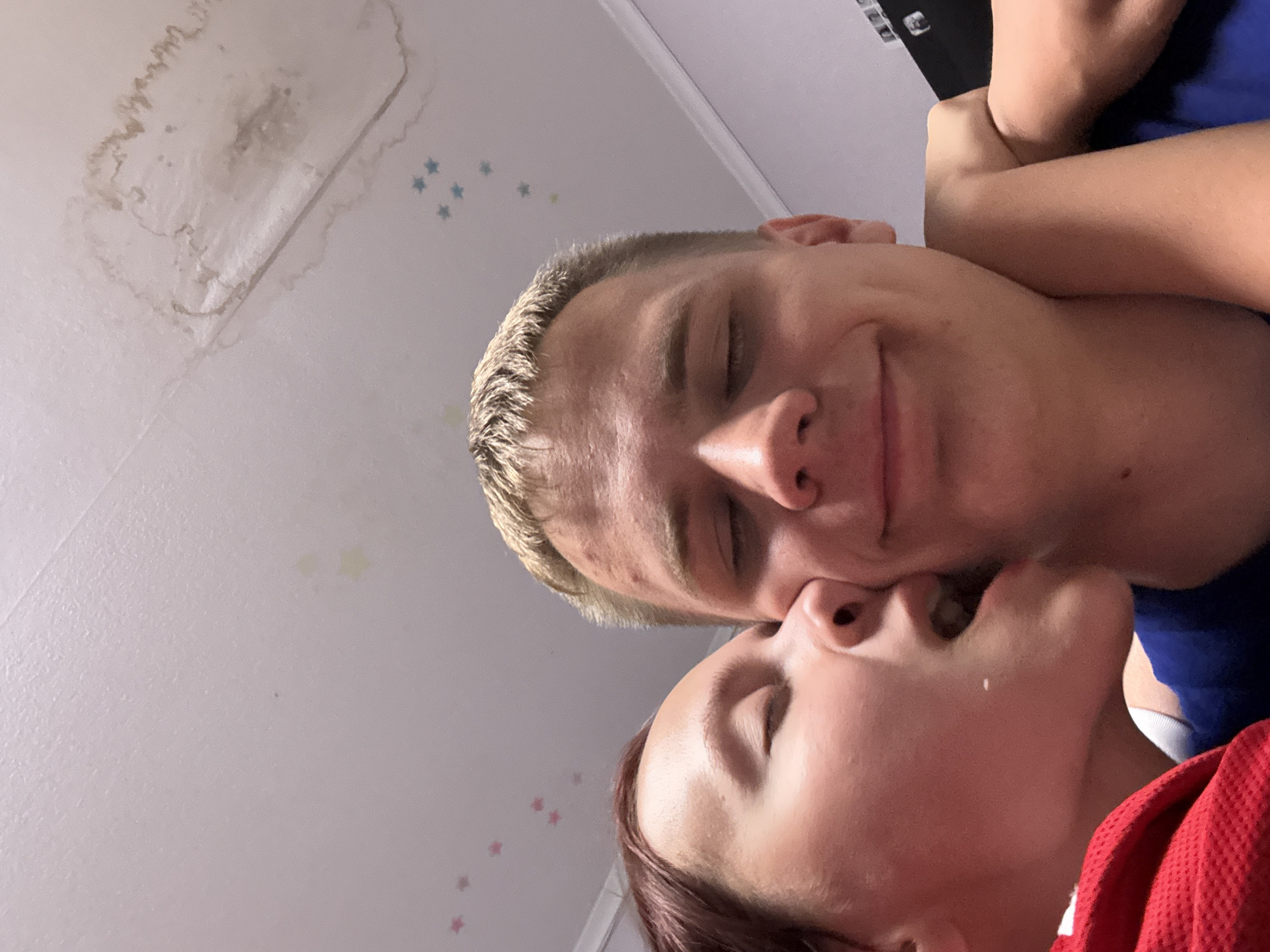 Two people are shown in a close-up, tilted selfie. The person on the left has blonde hair, closed eyes, and a slight smile. The person on the right has auburn hair, closed eyes, and is kissing the first person. In the background, a ceiling is visible with what appears to be a water stain and star-shaped decals. The location is in Прикамский, Russia.