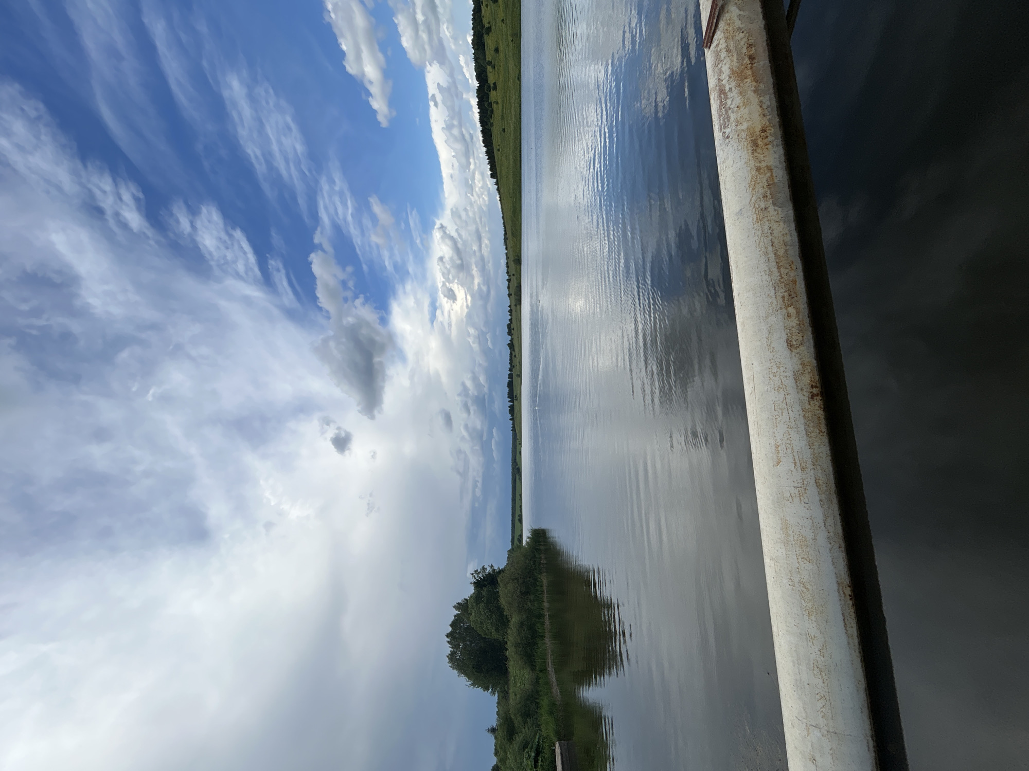 The image depicts a scenic view of a lake in Верх-Рождество, Russia. The foreground is dominated by a weathered, white pipe that stretches horizontally across the lower portion of the frame, partially submerged in the dark water. The lake itself is calm, reflecting the sky and the surrounding landscape. On the left side of the lake, a small island covered in dense green trees and bushes is visible. The distant shoreline features rolling green hills dotted with trees. The sky above is a dynamic mix of bright blue patches and dramatic white and gray clouds, suggesting a variable weather day. No people are present in the image, and no actions or interactions are captured. The overall environment is one of natural beauty and tranquility.