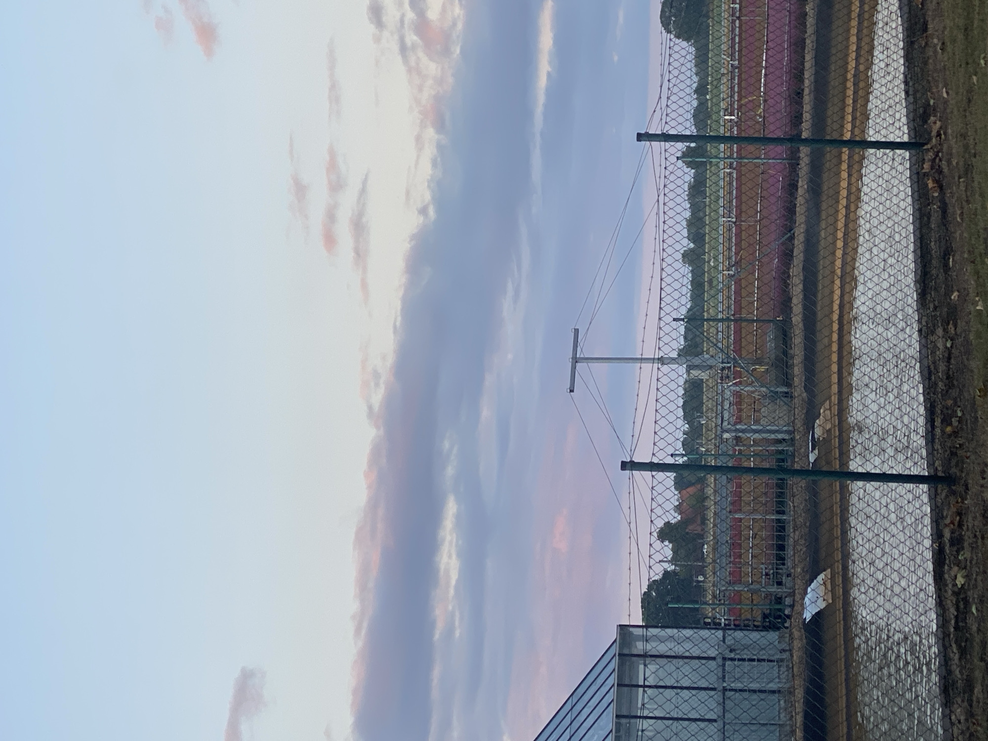The image depicts a cloudy sky with hues of pink and blue over an outdoor scene in Schwarme, Germany. In the foreground, a chain-link fence encloses a shallow body of murky water. Beyond the fence, a building with a glass facade and a metal roof is visible on the left. A metal pole with wires stretches across the middle ground. Further back, several long, rectangular structures, possibly tanks or containers, are lined up, some colored in shades of pink and orange. A line of trees forms the horizon, with a few buildings with red roofs nestled amongst them. The overall impression is of an industrial or agricultural area under a twilight sky. There are no people or animals visible in the image.