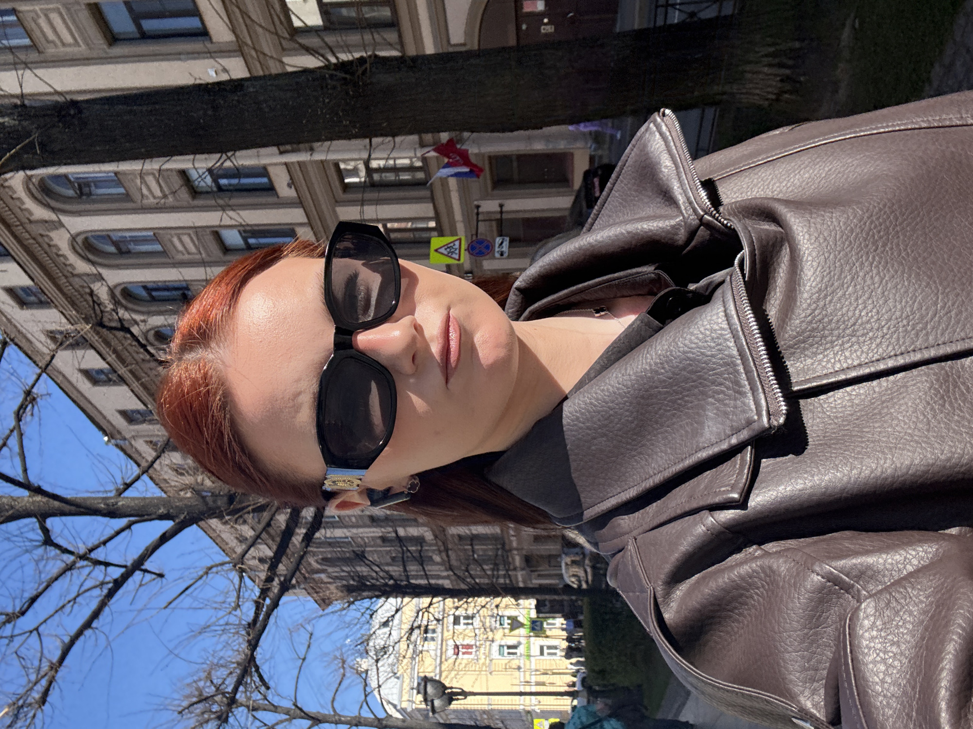 A woman with red hair wearing sunglasses and a brown leather jacket stands on the street of Saint Petersburg, Russia. Behind her is a tree and several buildings. A road sign and some flags are also visible. The day appears to be sunny.
