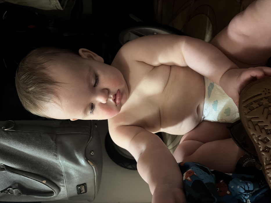 A baby is asleep in a stroller. The infant is shirtless, wearing a diaper with a light blue and white pattern. The baby is lying on a blue blanket with foxes. A gray diaper bag is visible to the left, with a stroller wheel and a brown shoe in the background. The baby's face is turned slightly to the left, with its eyes closed.