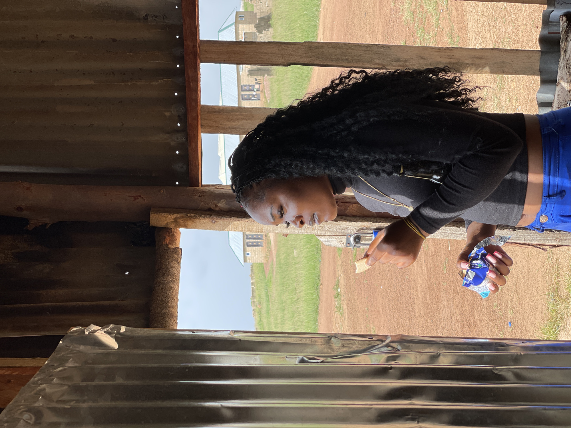 In Durumi, Nigeria, a young Black woman stands in a rudimentary structure with a corrugated metal wall to her left. She is looking to her left, her face in profile, with a piece of food in her right hand near her mouth and a snack wrapper in her left hand. She wears a black turtleneck shirt and bright blue pants. In the background, two simple stone buildings with blue roofs are visible across a grassy field under a cloudy sky. The ground is reddish-brown dirt.