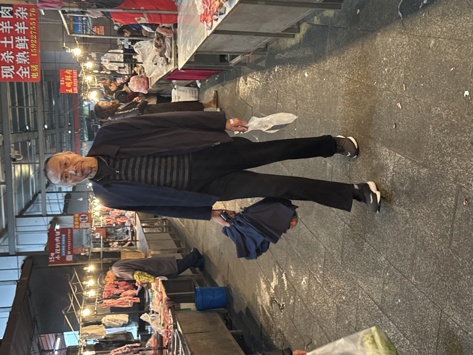 The image depicts an indoor meat market scene in Jiangjin, China. In the foreground, an East Asian male, appearing to be elderly, walks directly toward the viewer with a neutral expression. He has short, graying hair and is wearing a dark blue zip-up jacket over a horizontal striped shirt (dark gray and light gray/white), dark trousers, and dark athletic shoes with white soles. He carries a closed, dark blue umbrella in his left hand and a translucent white plastic bag containing an indistinct item in his right hand.
The market aisle features tiled flooring that appears damp and has scattered debris, including what resembles cigarette butts. On both sides of the aisle, numerous stalls are visible under artificial lighting. To the left, a vendor, clad in a smock with yellow sleeves, is actively preparing meat at a metal counter, with various cuts of raw meat hanging above and displayed on the counter. A blue bucket is positioned near his stall. A red sign above this area reads "15号 小双的肉铺" (No. 15, Xiaoshuang's Meat Shop) with a phone number "19909399998".
To the right, similar meat stalls are present with more raw meat on display and hanging. Other individuals are visible further down the aisle, some appearing to be customers or vendors. Signs in Chinese characters are prominently displayed, including one at the top right advertising "现杀土羊肉 全熟鲜羊杂" (Freshly slaughtered local lamb, fully cooked lamb offal) with a phone number "15922755176". Another sign reads "53. 王明鲜肉" (No. 53, Wang Ming Fresh Meat). The overall environment is illuminated by a mix of overhead industrial lighting and warm-toned individual stall lights, characteristic of an active, bustling market.