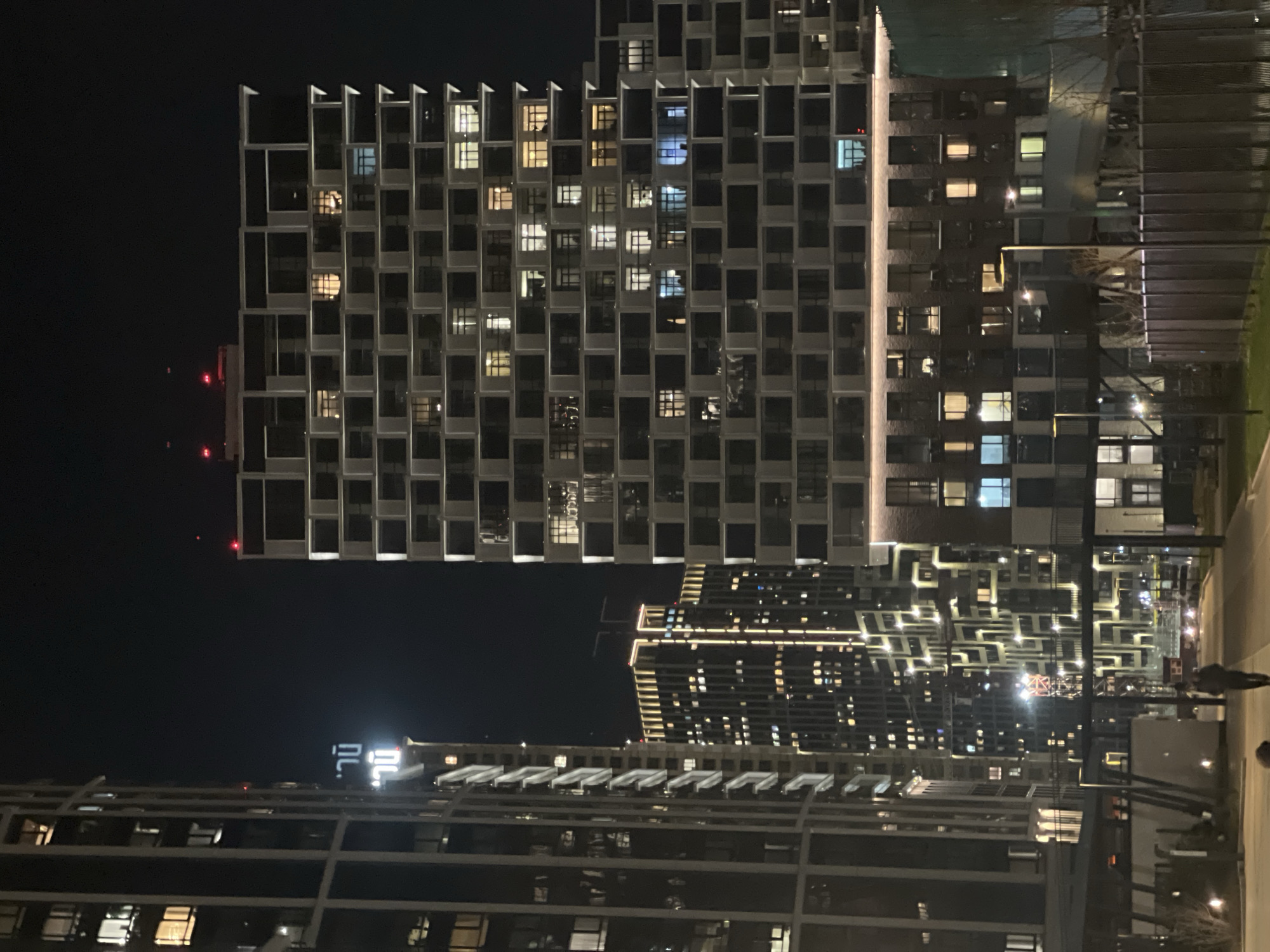 The image depicts a modern urban cityscape at night. The scene is dominated by several large, illuminated buildings under a dark sky.
In the foreground and slightly to the left, a tall, multi-story building features a distinctive grid-like facade with numerous recessed windows, many of which are lit, casting warm and cool glows. Red warning lights are visible on the top of this building and another structure in the distance. To the right of this building, another structure appears less defined, but also displays numerous lighted windows.
Further back and to the right, a complex of buildings showcases an intricate pattern of bright, linear lights along its facades, creating a striking visual effect against the night. On a distant building, glowing, stylized text, possibly Cyrillic characters "ПЛ." (PL.), is faintly visible.
At the bottom right of the image, what appears to be a street or paved pathway is illuminated by streetlights, and a few blurry figures of people can be seen walking, suggesting pedestrian activity. The overall impression is one of a bustling, modern city quieted by the late hour, with architecture and artificial light defining the environment. The clear night sky indicates no adverse weather conditions.
