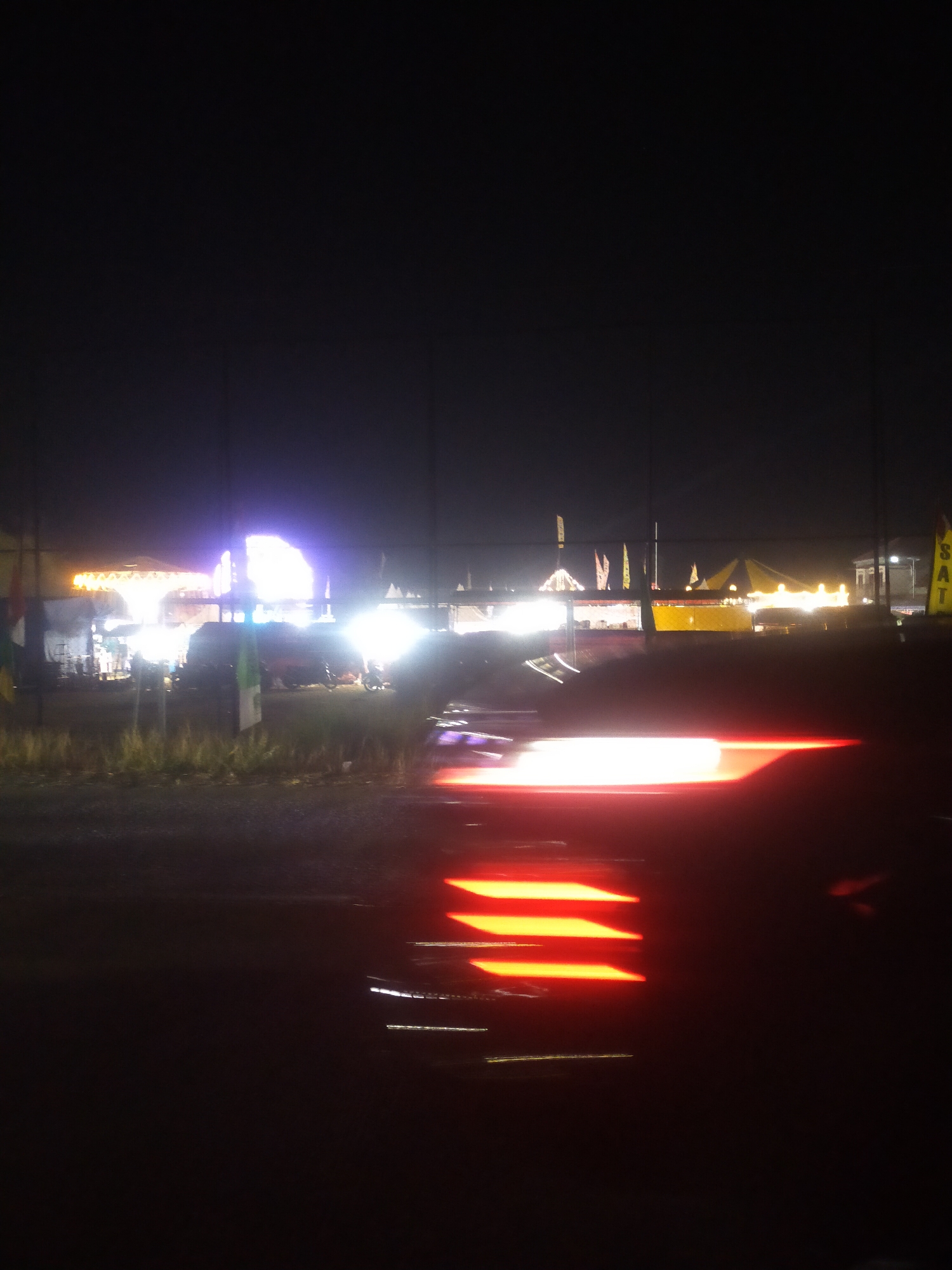 The image depicts a scene at night, likely a festival or market, in Rengaspendawa, Indonesia. The foreground is dominated by the blurred red and white light trails of a moving vehicle, suggesting a long exposure photograph. In the midground, there are several tents and structures illuminated by bright lights, some appearing to be market stalls or event venues. Flags are visible, indicating a celebratory or organized event. The background is dark, with only faint hints of distant lights. The overall impression is one of a lively night scene, possibly a temporary gathering, captured with motion blur due to the vehicle's movement.