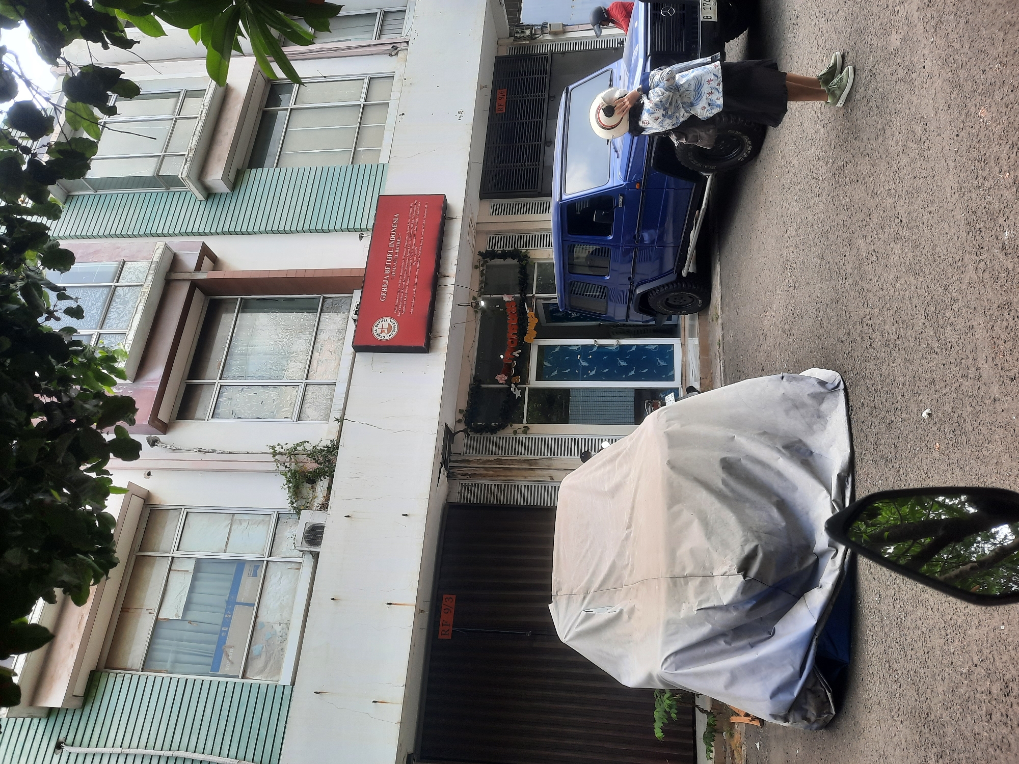 The image shows a street scene in front of a multi-story building in North Jakarta, Indonesia. The building has a light-colored facade with several windows, some of which are covered or partially obscured. A red sign above the entrance reads "GEREJA BETHEL INDONESIA" with additional text in a smaller font below.
In front of the building, a blue Mercedes-Benz G-Wagen is parked. To the left of the G-Wagen, a car is covered with a light-colored tarp. A woman wearing a floral shirt, black skirt, and a hat is standing in front of the G-Wagen, facing the building and holding what appears to be a cell phone to her ear. To the right of the G-Wagen, another person wearing a red shirt and helmet is partially visible, possibly on a motorcycle.
The street is paved with asphalt, and there are some trees and foliage in the foreground and around the building. The overall impression is that of a commercial or institutional building in an urban environment.